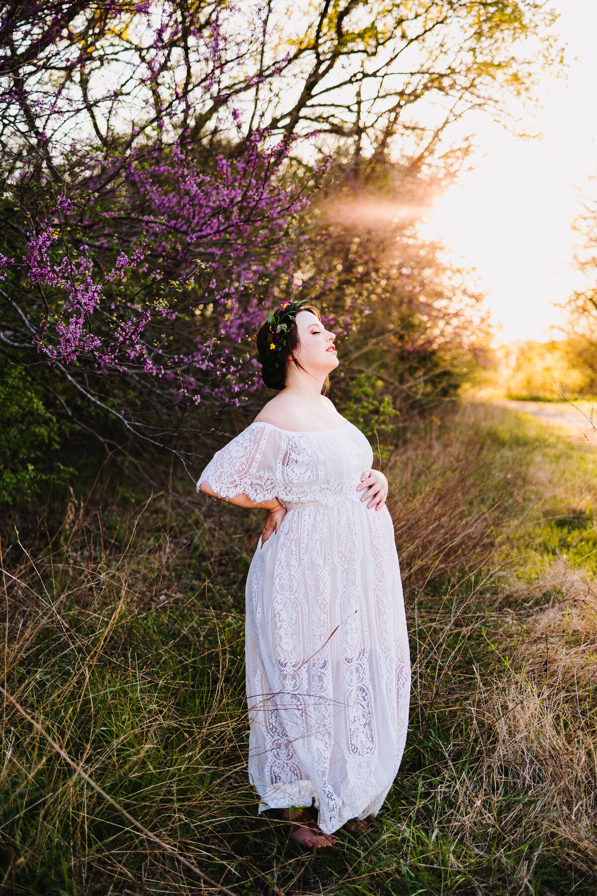 Photo of a pregnant woman in a garden with purple flowers. The woman has a long white dress and a flower headband with different colors