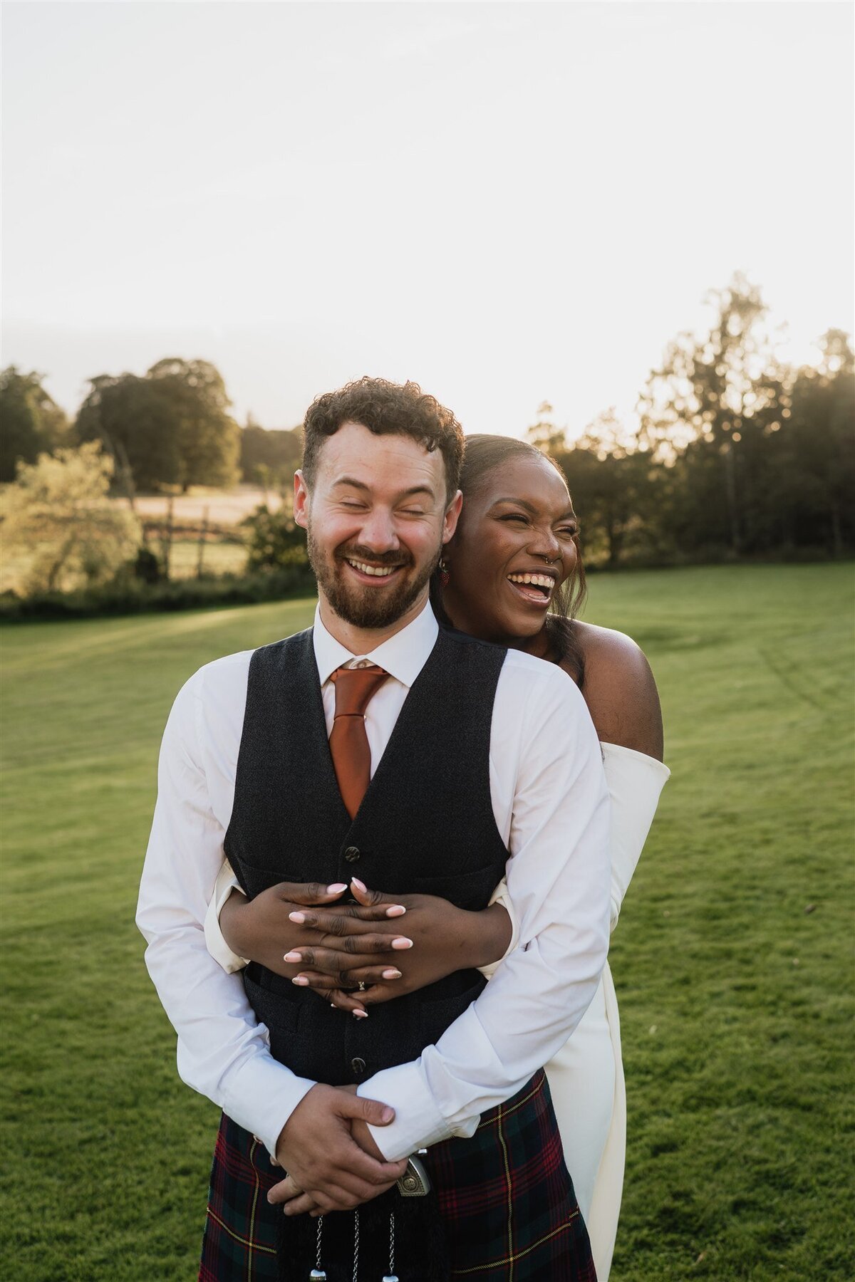 A bride wraps her arms around her husband from behind and they laugh together.