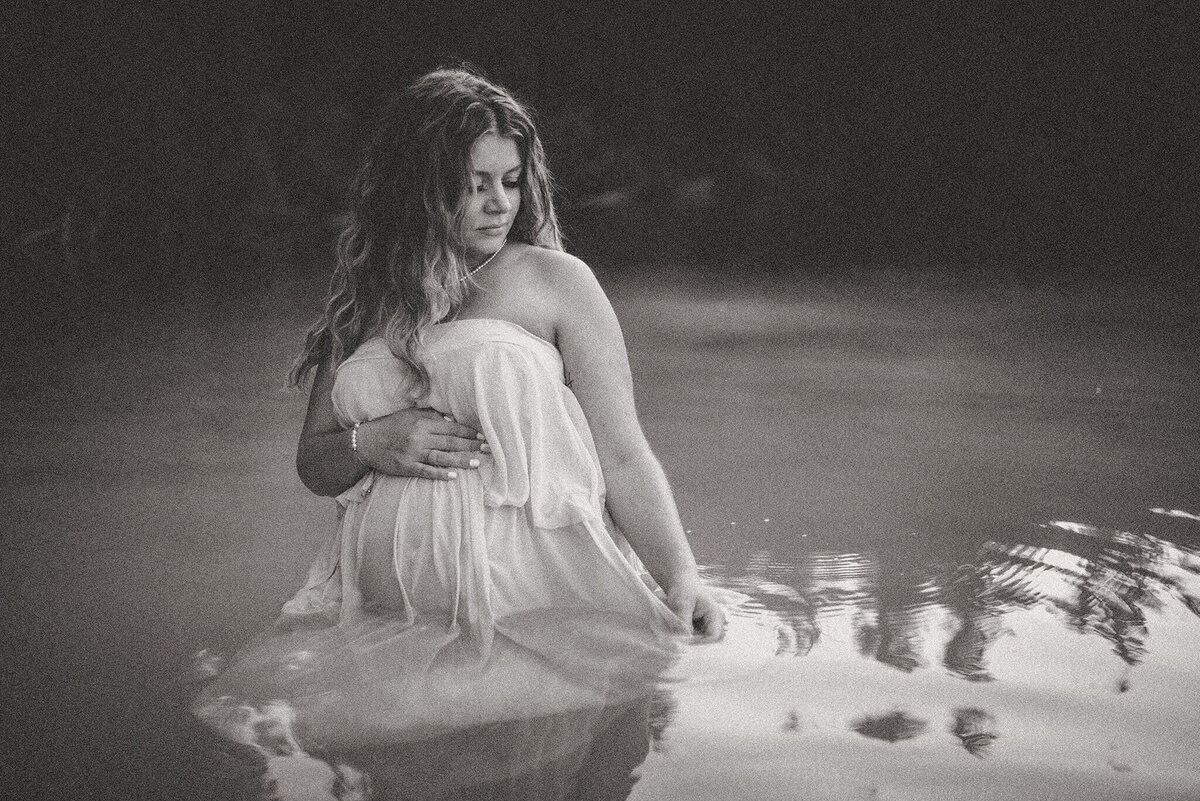 Black and white portrait of a pregnant woman in water. She is wearing a flowing dress.