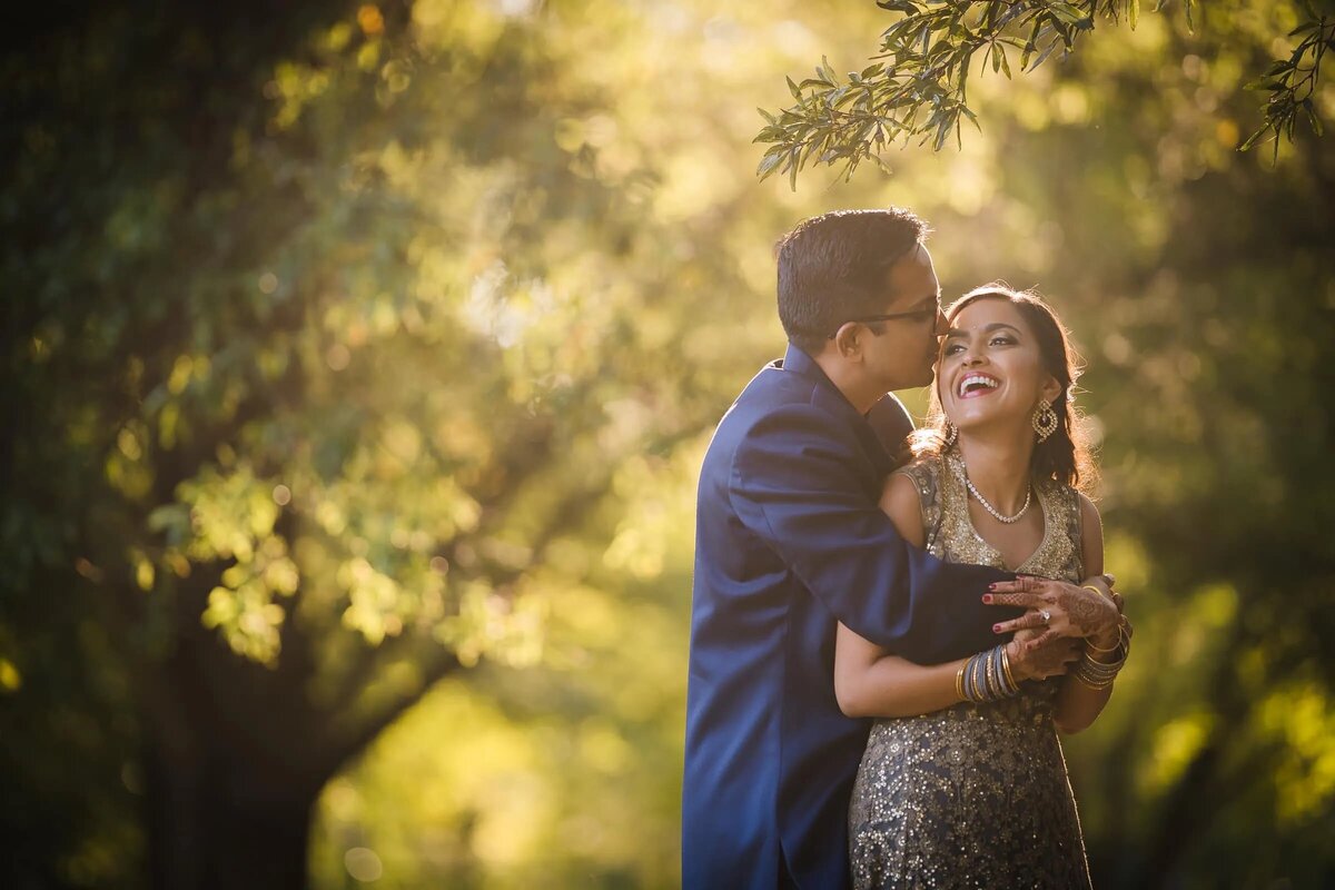 A couple shares a laugh in a sunlit garden, the groom's blue suit and the bride's shimmering dress glowing in the natural light.
