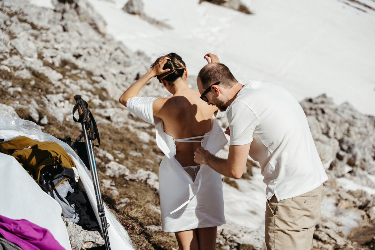 Groom helping bride into wedding dress on snowy mountain trail