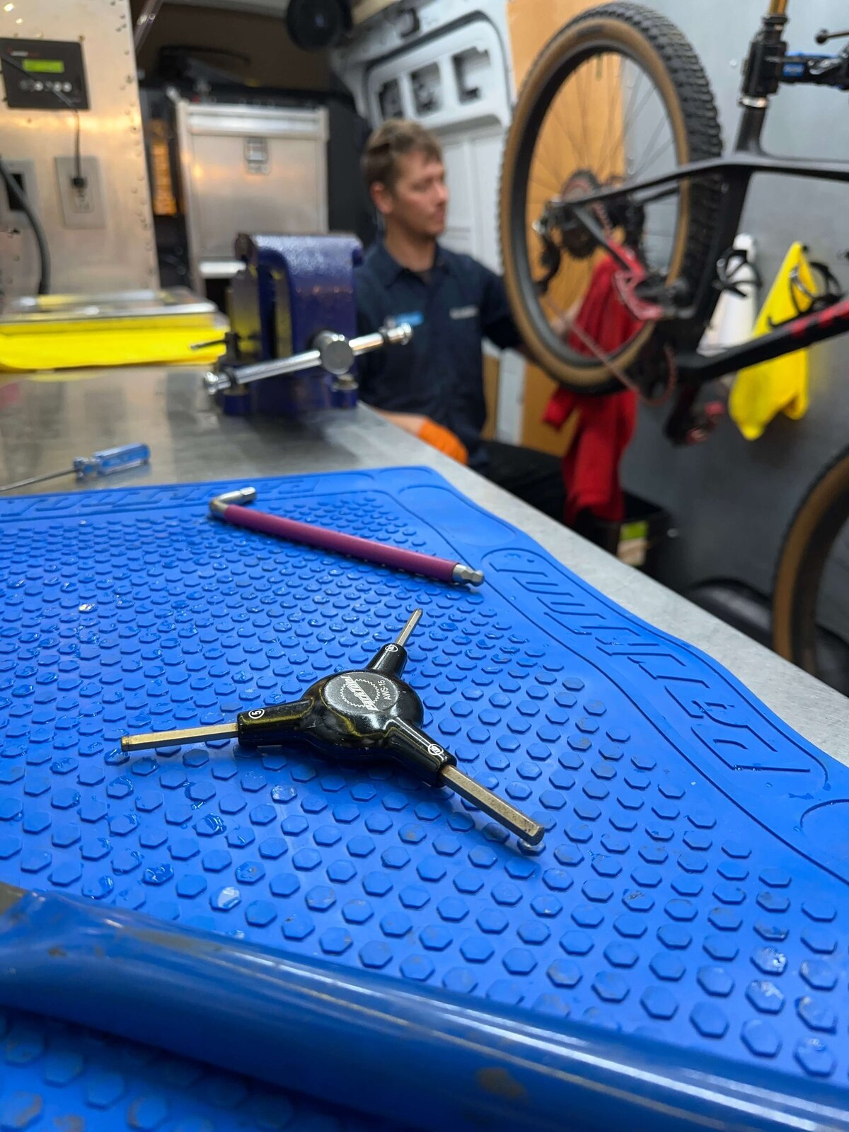 A bike repair station with a blue mat in the foreground, featuring a multi-tool and a pen. A mechanic in the background works on a mountain bike secured in a repair stand. Various tools and equipment are visible