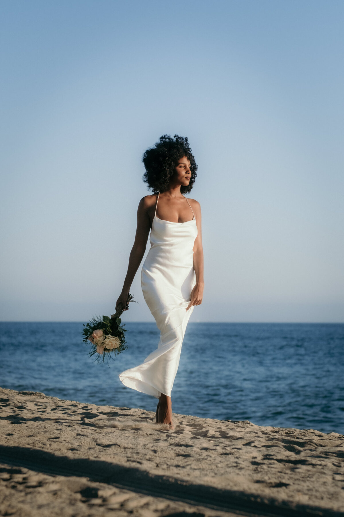 With the blue sea in the back and her bridal bouquet in her hand, the bride walks over the beach.
