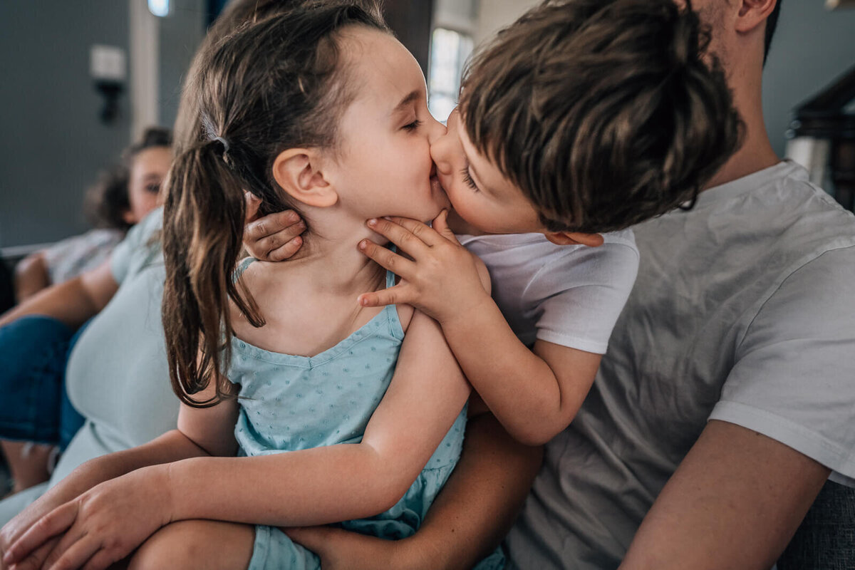 Two young siblings kiss during an in home maternity photography session.