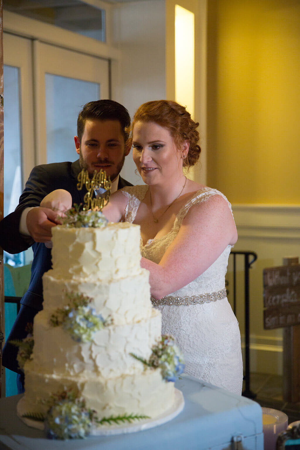 A bride and groom are cutting a three-tiered white wedding cake decorated with flowers. The bride, in a lace dress, and the groom, in a dark suit, are both holding the cake knife together.