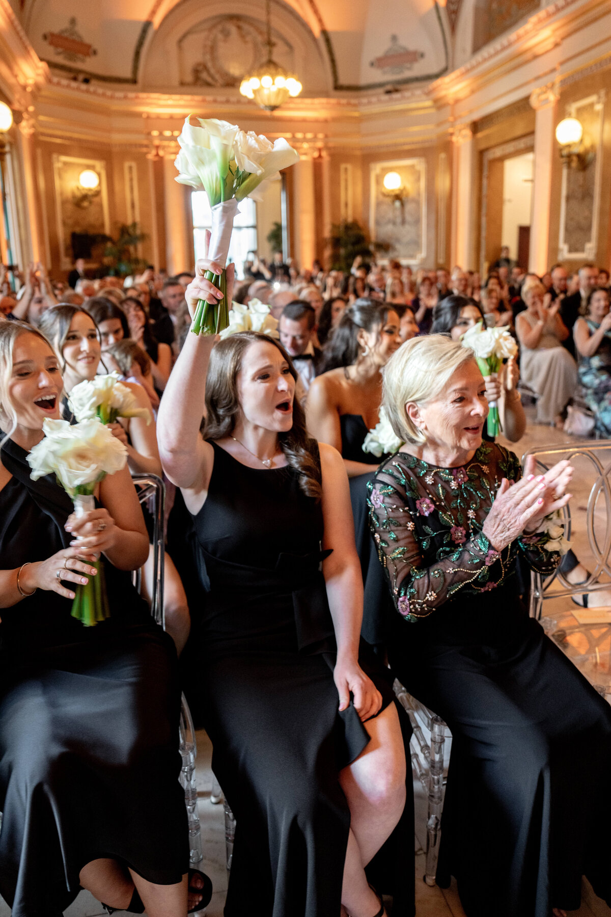 A joyous group of people seated in an elegant hall. A woman in a black dress holds a bouquet of white flowers aloft, smiling, while others nearby clap and cheer. The ornate room is well-lit with chandeliers.