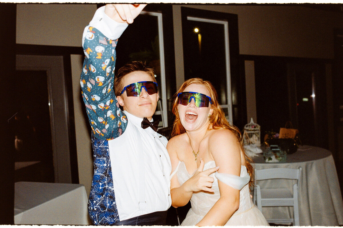 Bride and groom, wearing sunglasses, laugh at the camera on the dance floor