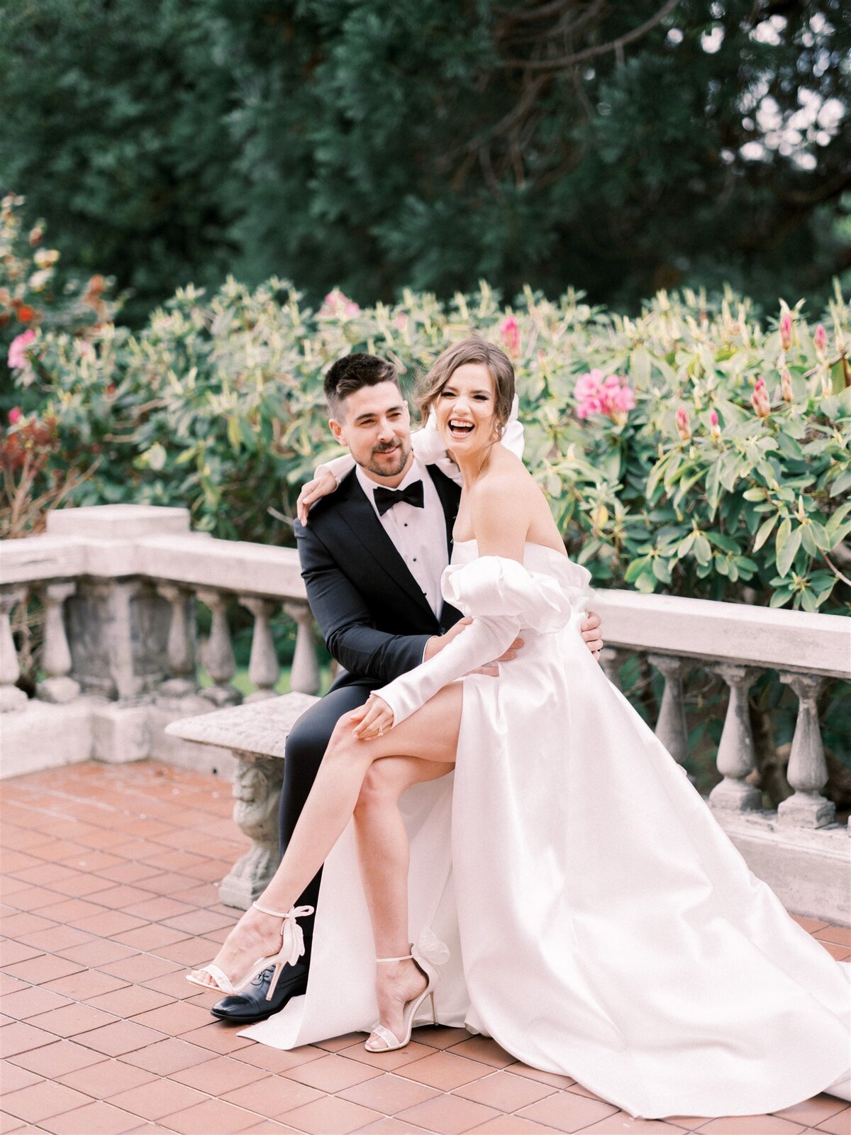 A couple sits on a stone bench; the man is in a black suit and bow tie while the woman wears a white off-the-shoulder dress and heels. Green foliage and pink flowers are in the background, all beautifully arranged by Destination Wedding Planner Melissa Dawn Event Designs.