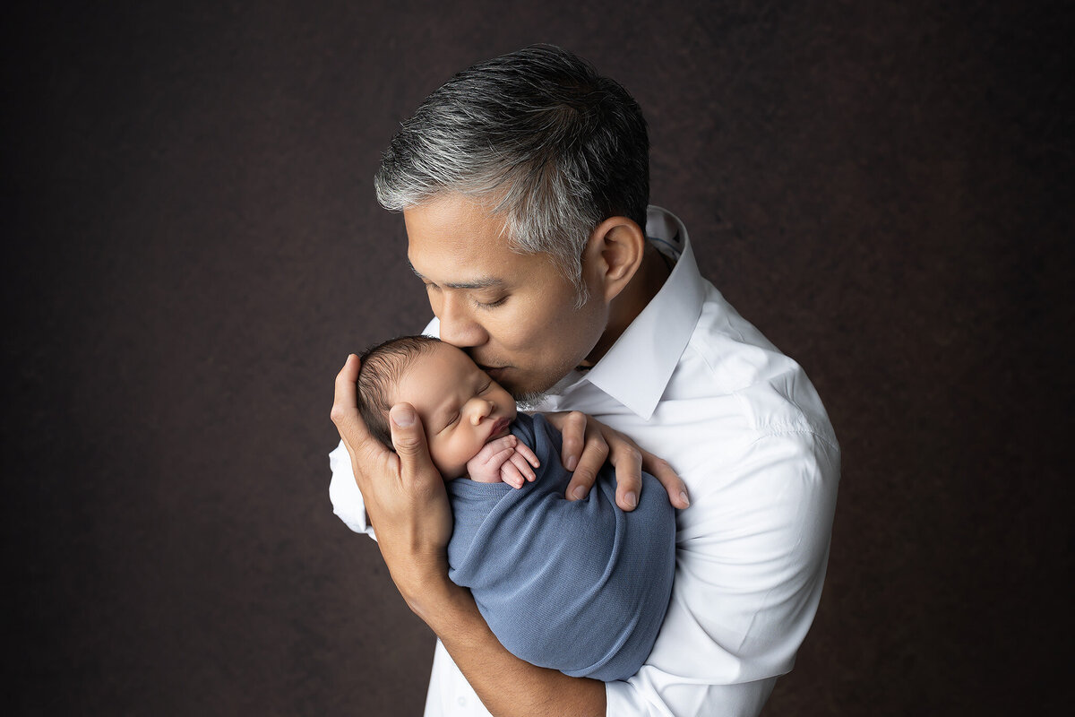 A father in a white shirt kisses his swaddled newborn's head, creating a peaceful and intimate moment of connection. Created in studio newborn session at Lindsay Walden Photography.