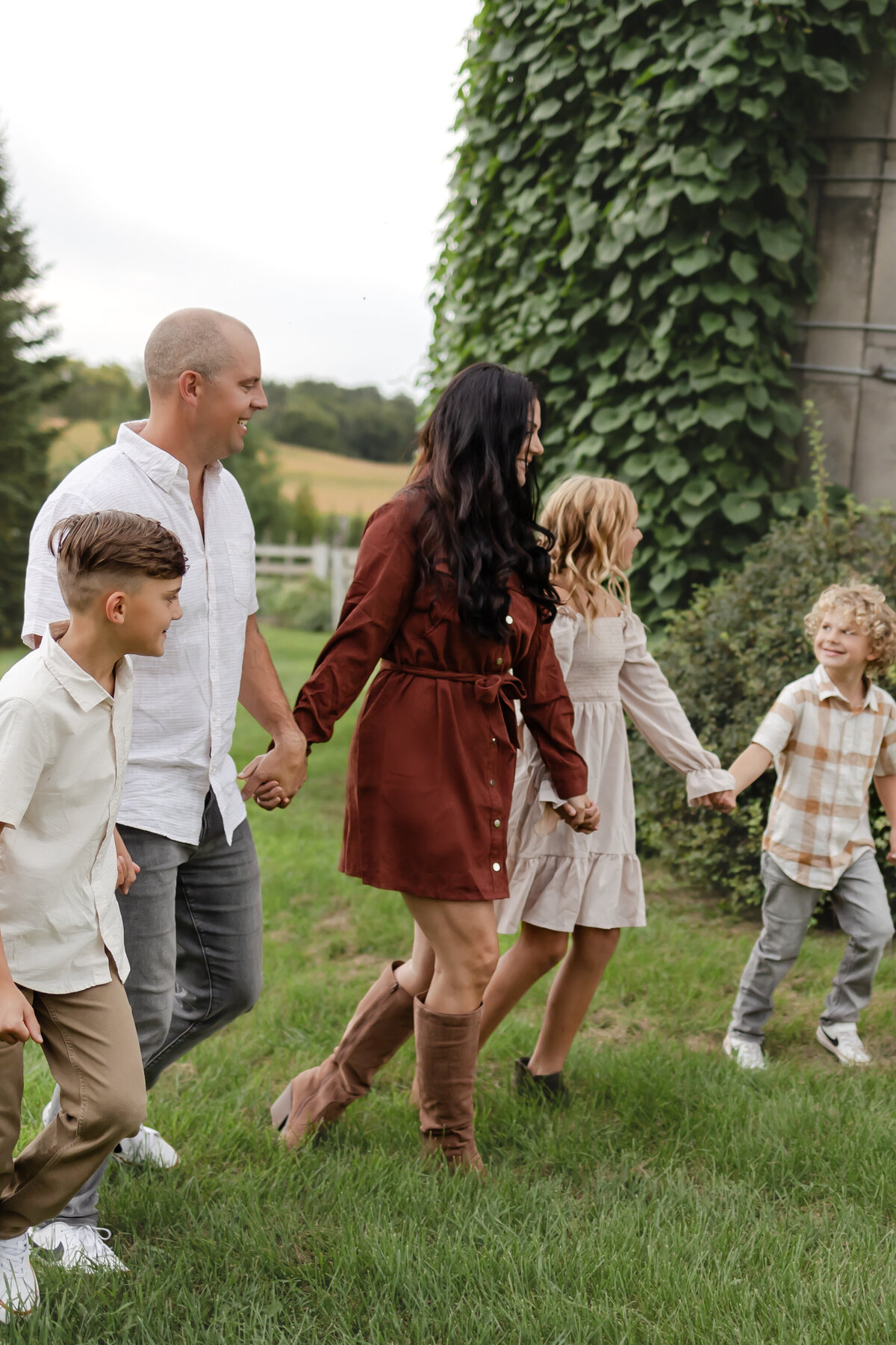 a family holds hands as they walk through green grass with cascading ivy down a silo at a farm, wearing fall attire