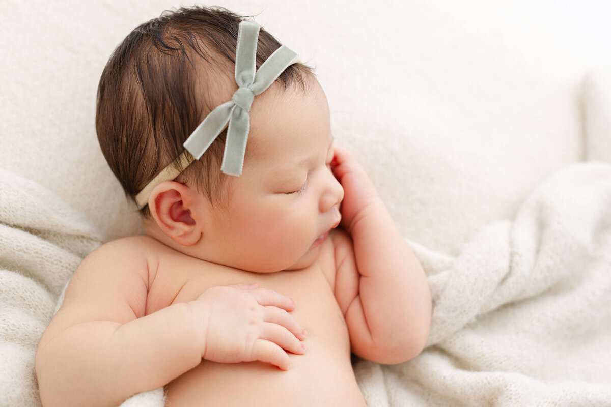 Baby sleeping with a green velvet bow on her head. One hand is resting on her chest while the other is up by her face.