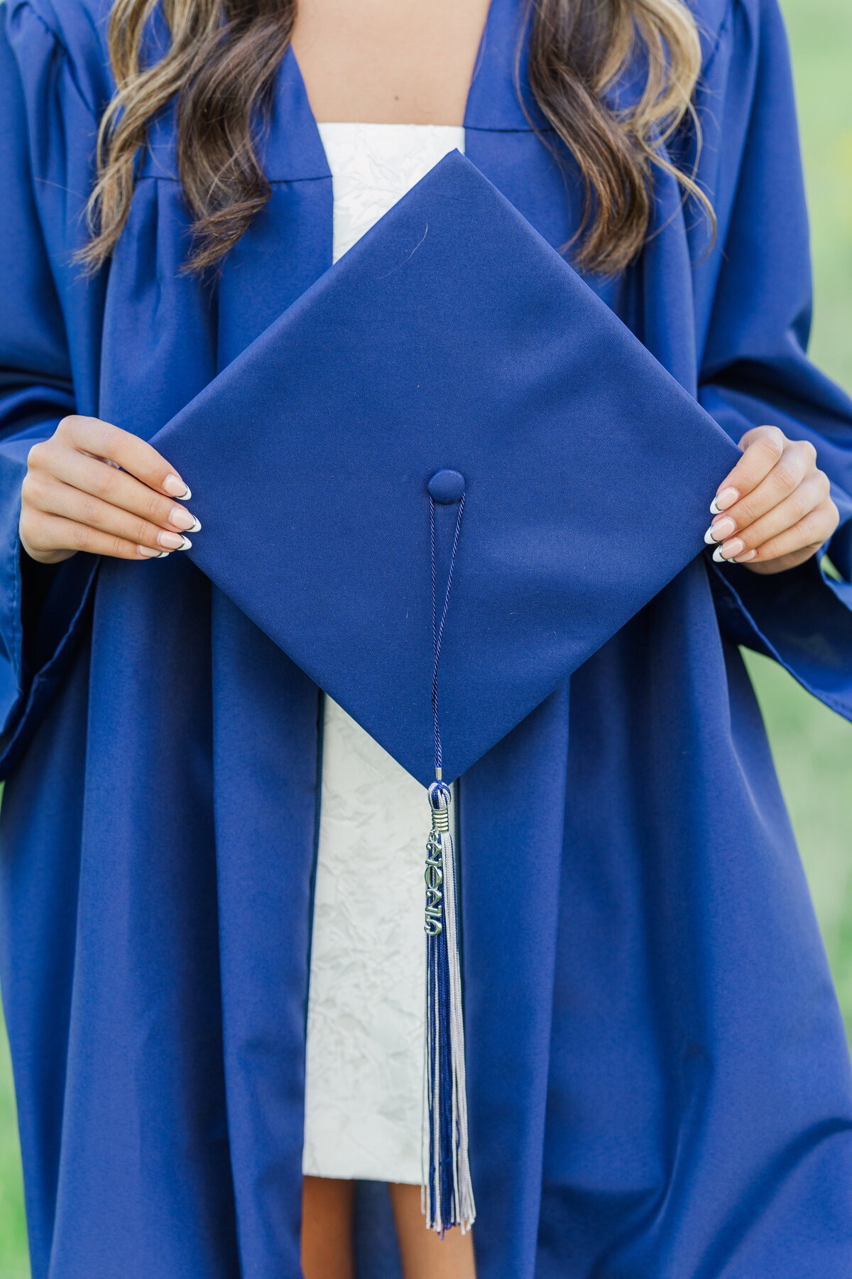 Close-up of a high school senior holding a navy graduation cap and tassel in a blue gown, photographed in a sunny field at Arbor Hills Nature Preserve in Plano, TX.