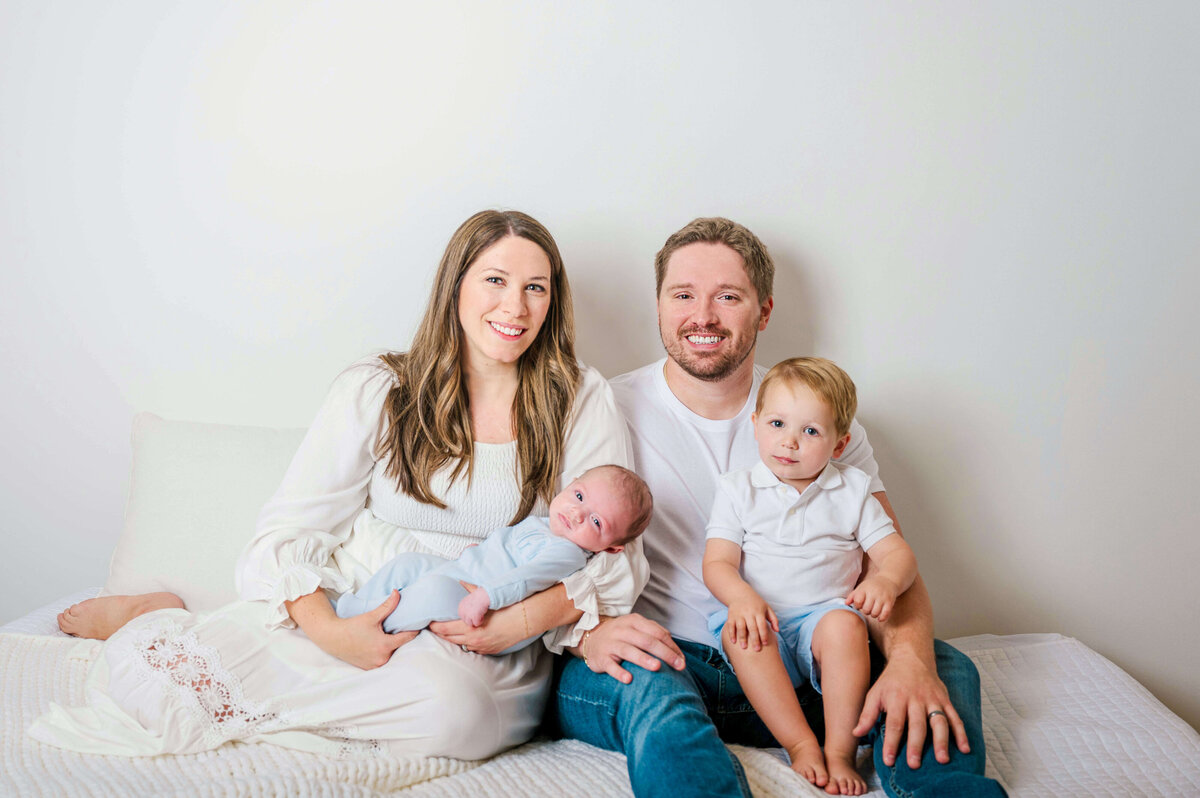 Cute young family, with mom, dad, young son, and a baby sitting on a bed and smiling at the camera. 
