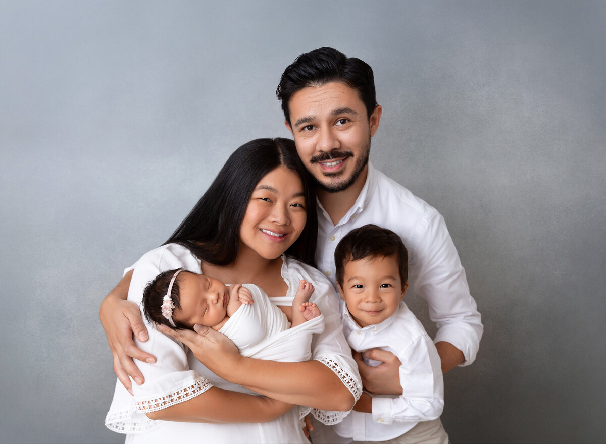 Family photo of new mom, dad, big brother, and baby sister. Baby is sleeping in mom's arms. Mom, Dad, and brother are smiling at the camera for photoshoot.