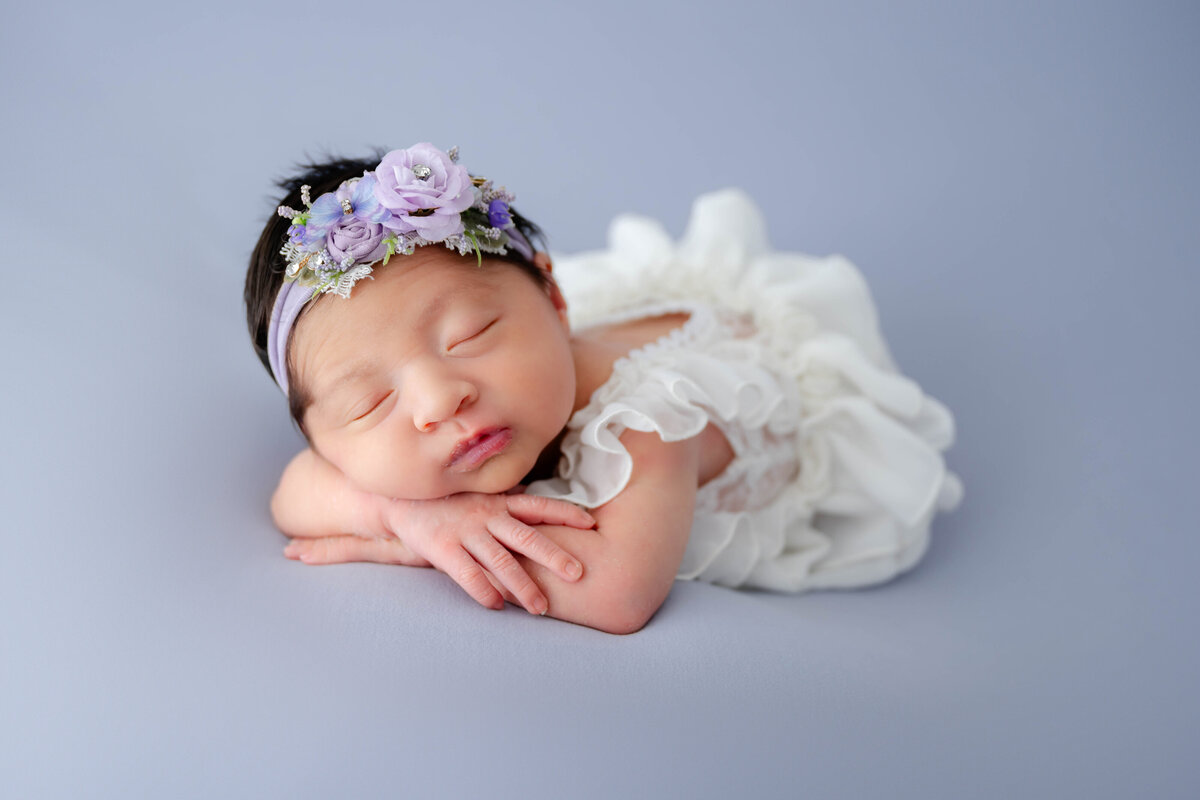 Newborn baby girl with chin on arms while laying on her belly and dressed in an all white dress.
