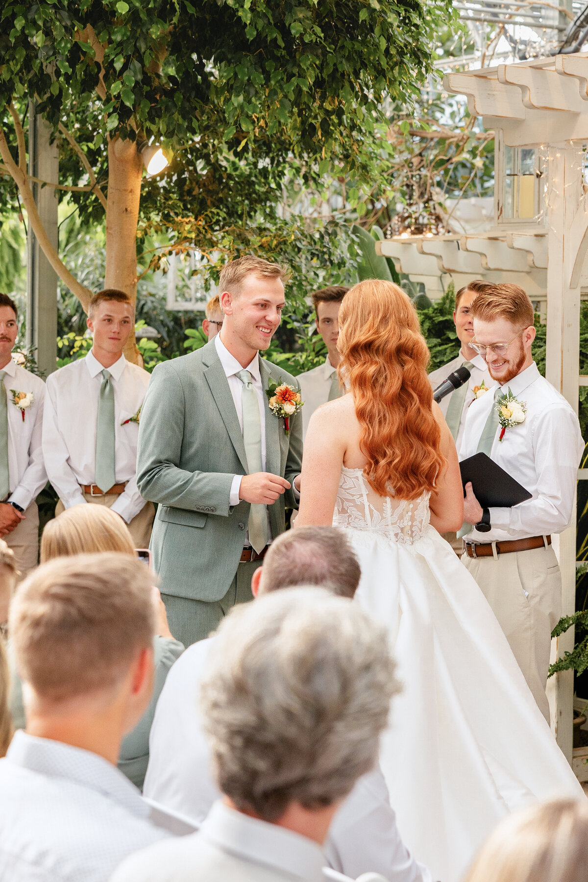 The groom, in a sage green suit, smiles as he exchanges vows with his bride, who wears a white gown with cascading curls, under twinkling lights and greenery.