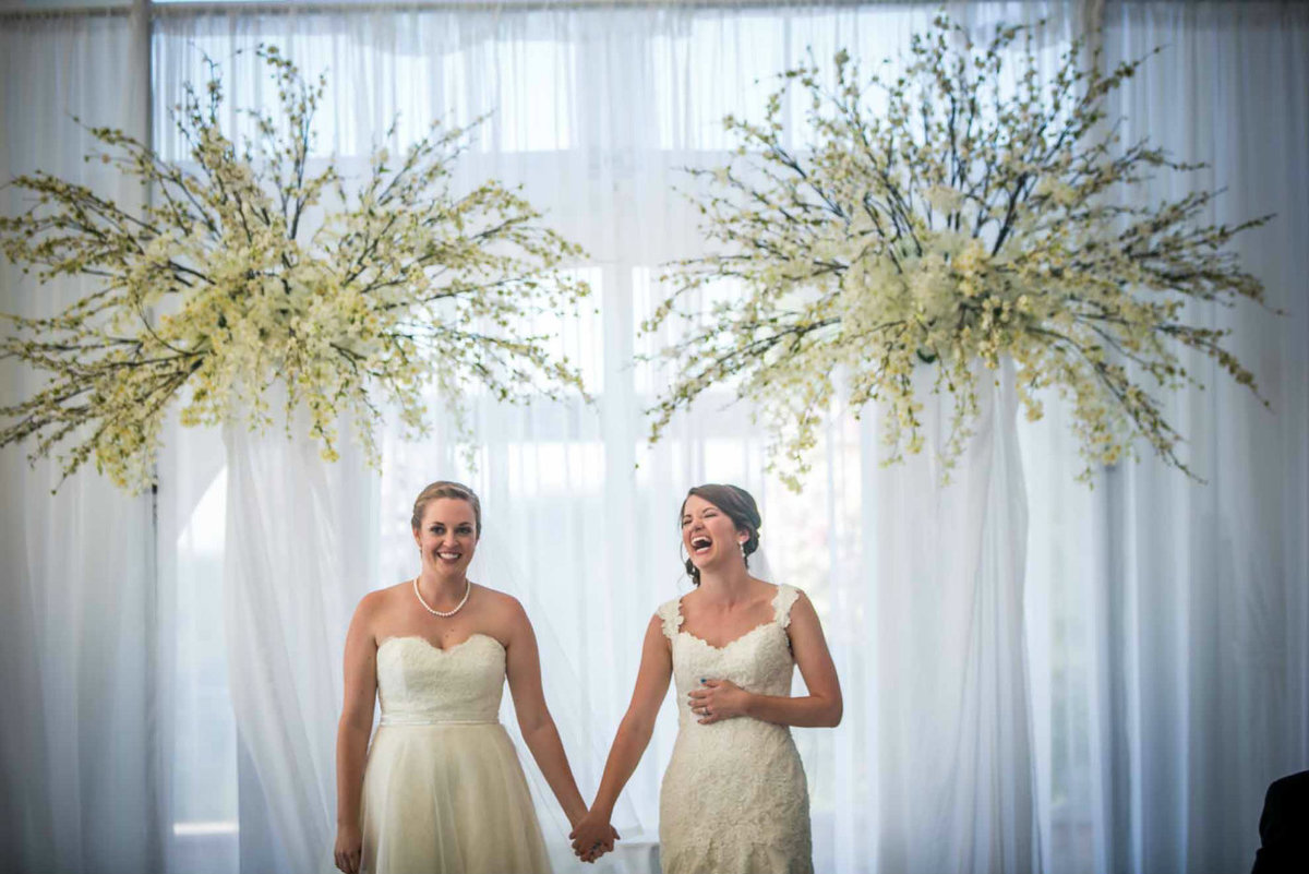 two brides in wedding dresses under tall white flowerarch