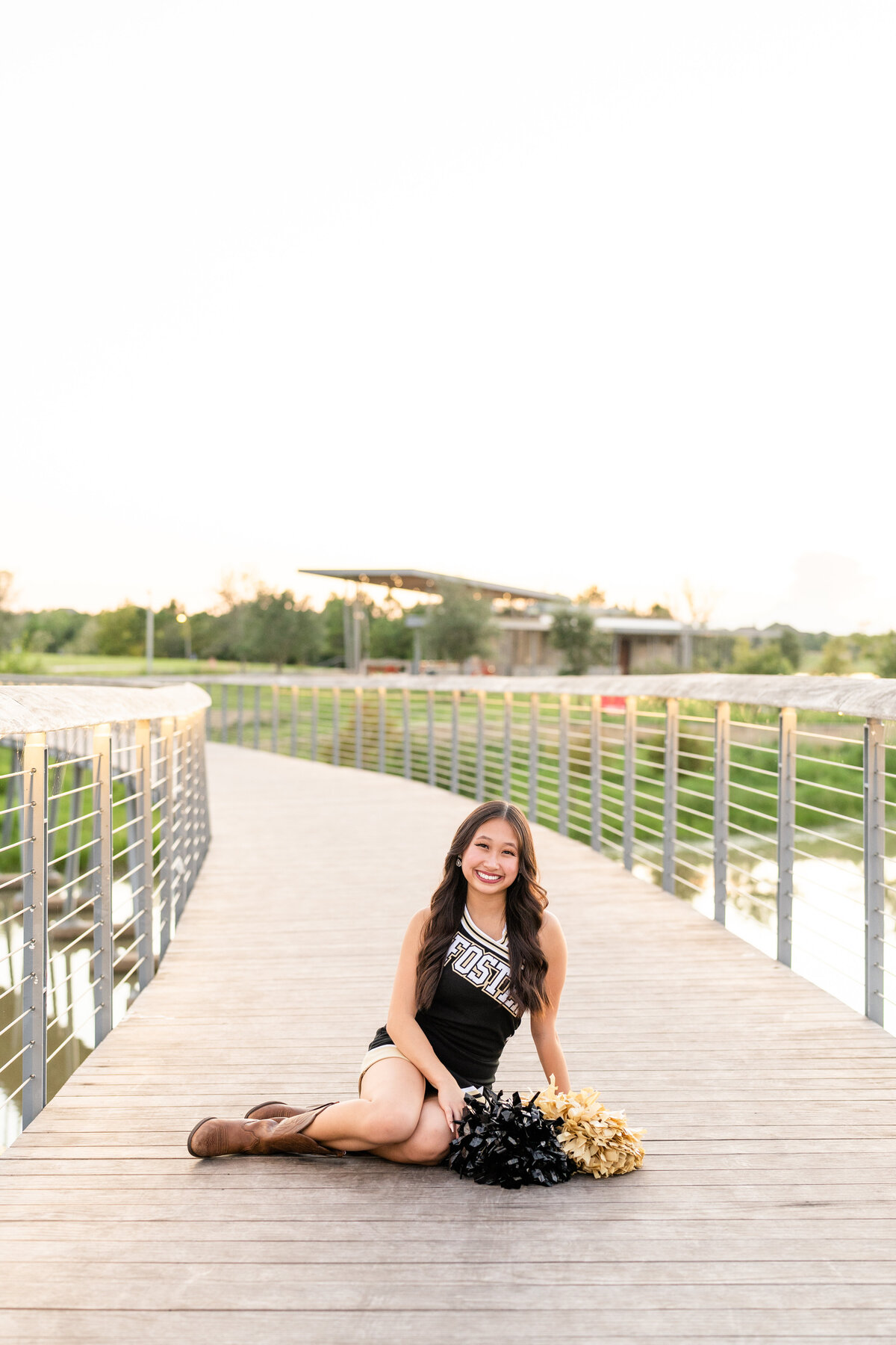High School senior girl sitting on bridge in cheerleading outfit with poms on ground and smiling at Josey Lake Park in Bridgeland