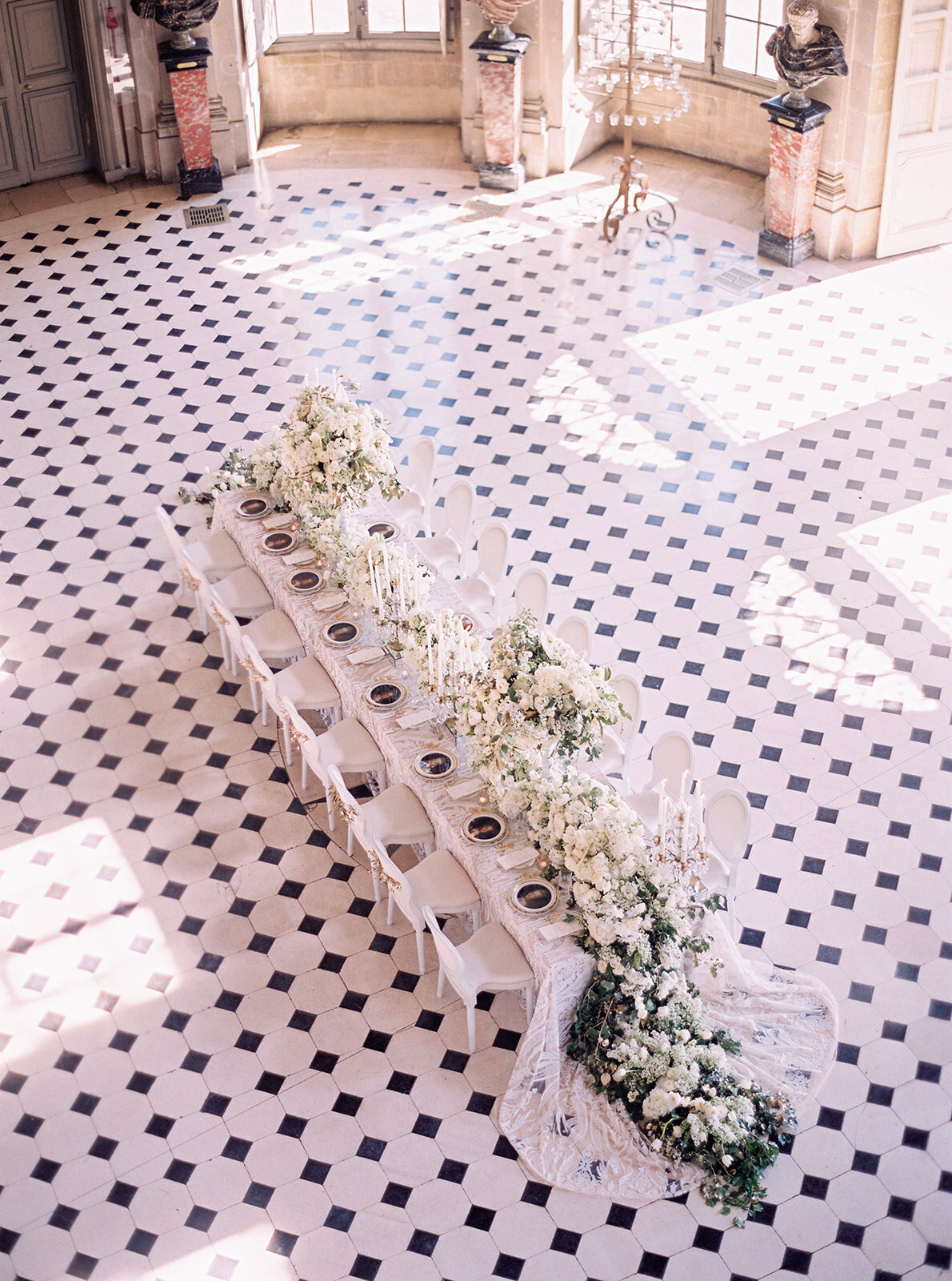 This stunning photograph was captured at the historic Chateau Vaux le Vicomte in Paris, France by the talented photographer Audrey FORA, with East West Films as the videographers. Known as one of the top destination wedding venues in France, this exquisite chateau provides the perfect setting for elegant events.  In the image, a long, elegant dining table is beautifully decorated with white flowers and greenery, creating a luxurious and formal ambiance. The grand room where the table is set features gorgeous black and white checkered flooring, adding a classic touch to the setting. Ornate columns and large windows frame the space, enhancing the historic grandeur of the venue.
