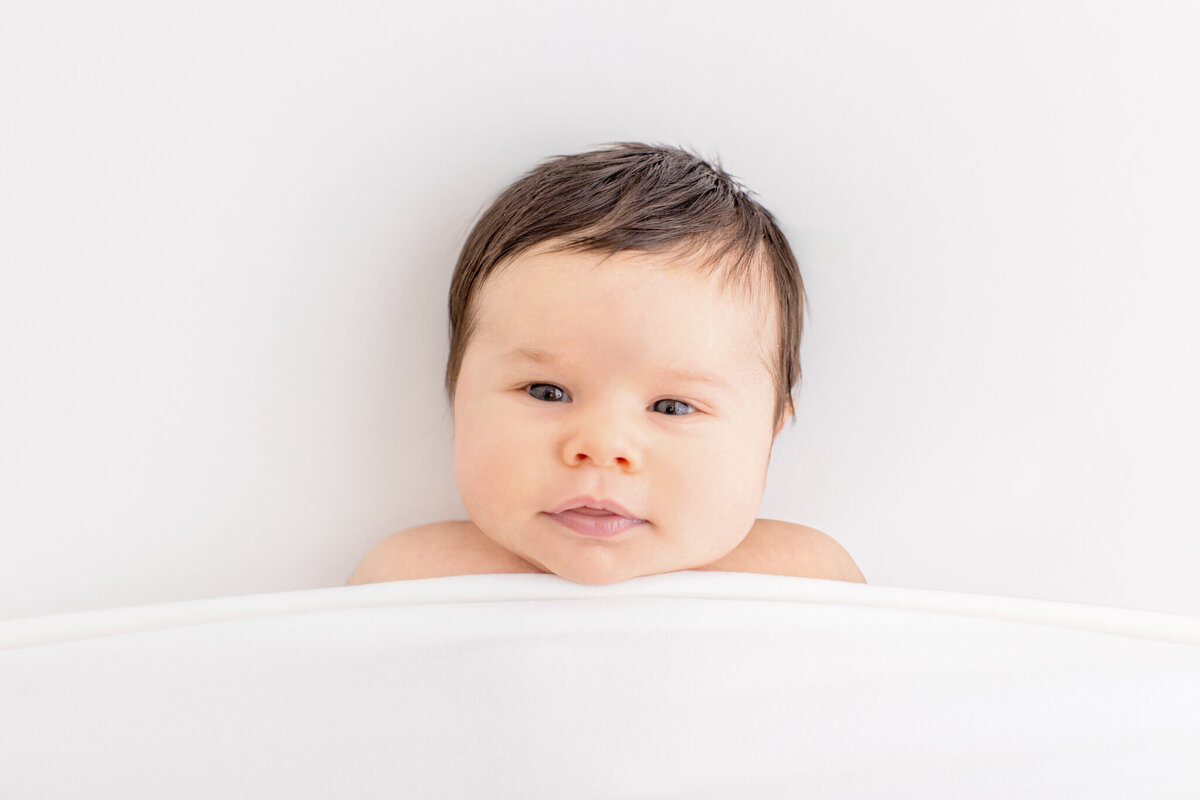 A baby with dark hair is partially covered by a white blanket, gazing upward with a calm expression against a soft white background.