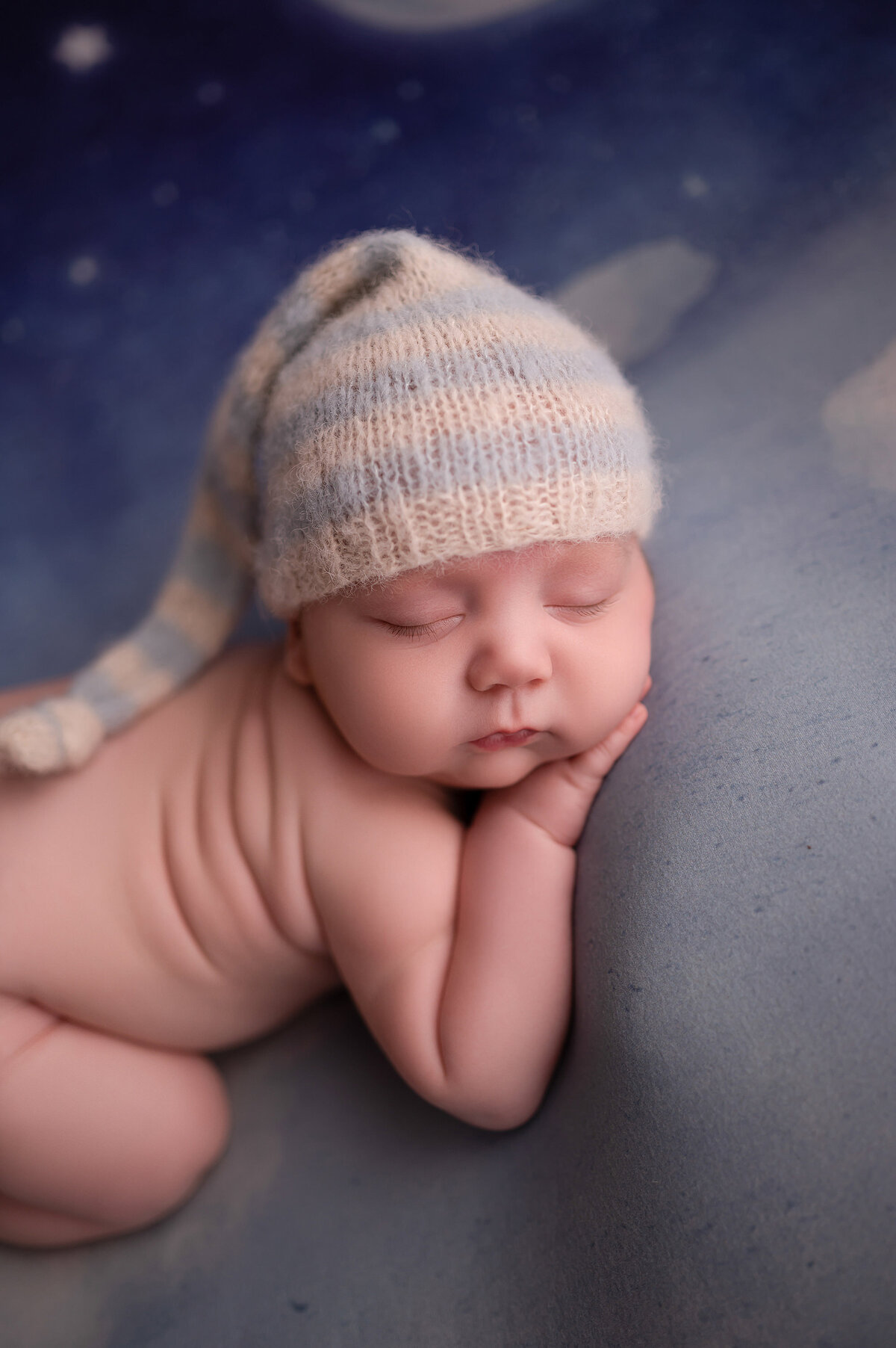 Portrait of a sleeping newborn baby wearing a striped stocking cap on a blue backdrop.