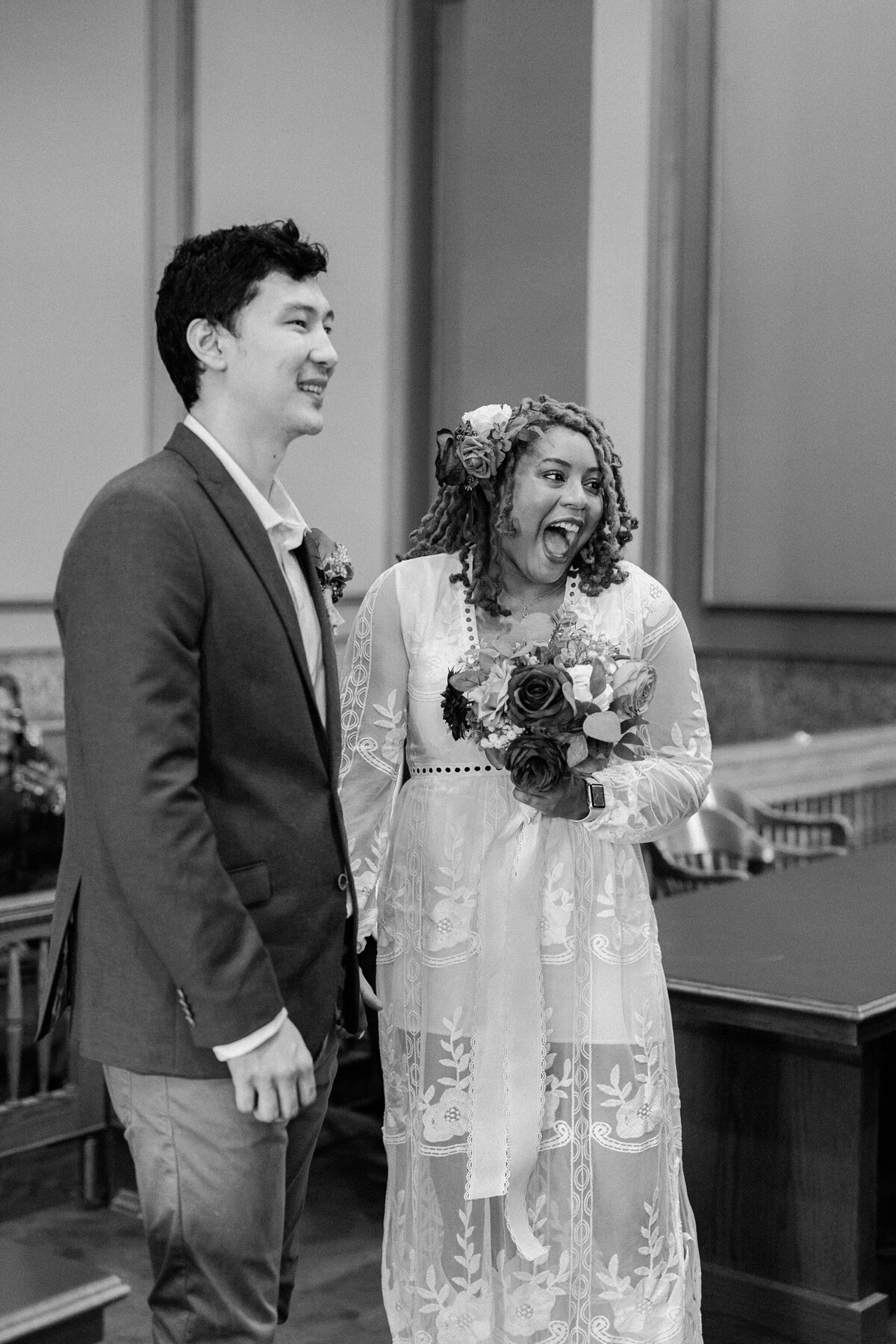 Black and white candid shot of a bride and groom during their wedding ceremony at the Tarrant County Courthouse in Fort Worth, Texas. The couple are both smiling and looking joyfully up at the judge who is out of frame. The groom is wearing a suit and boutonniere. The bride is wearing a long sleeve, intricate, white dress and is holding a bouquet.