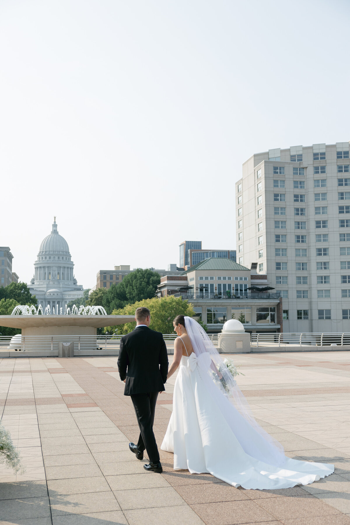 The bride and groom cheerfully exit their ceremony at The Madison Club, with the iconic Wisconsin State Capitol providing a beautiful backdrop to their joyful celebration.