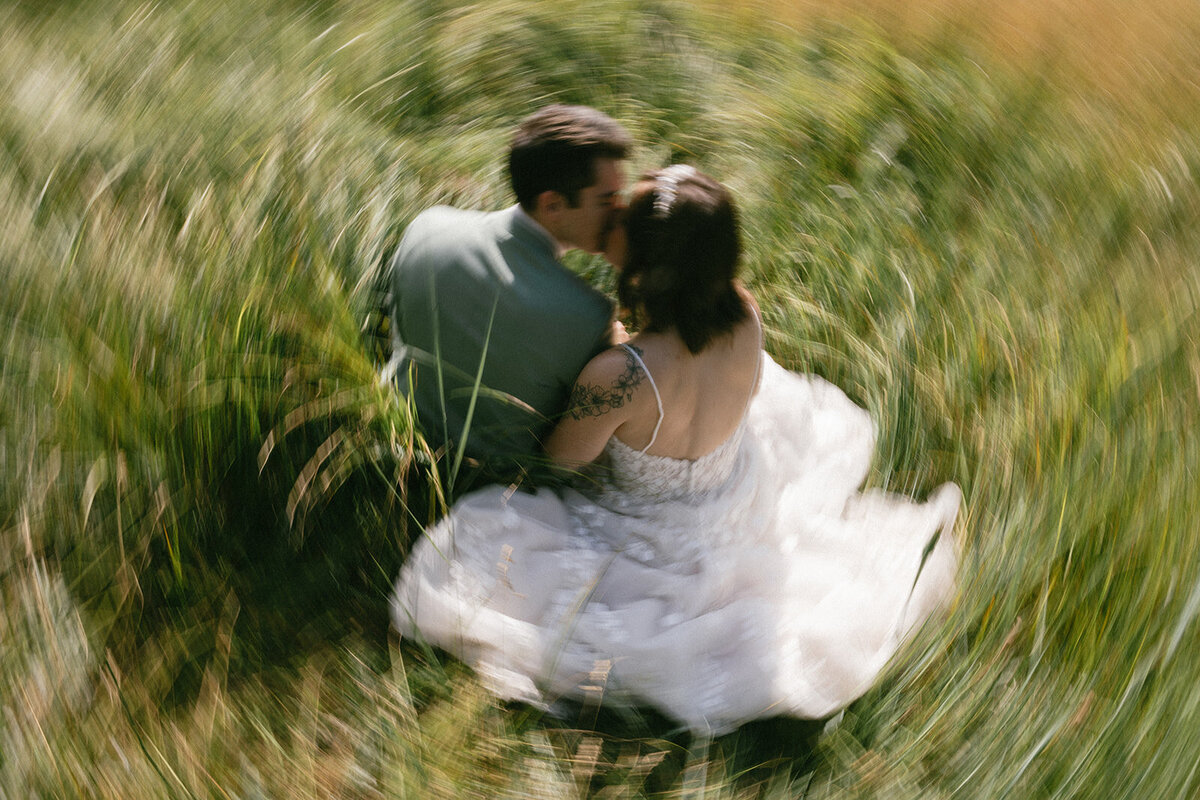 Elopement couple sits in a meadow while grass swirls around them on their wedding day.