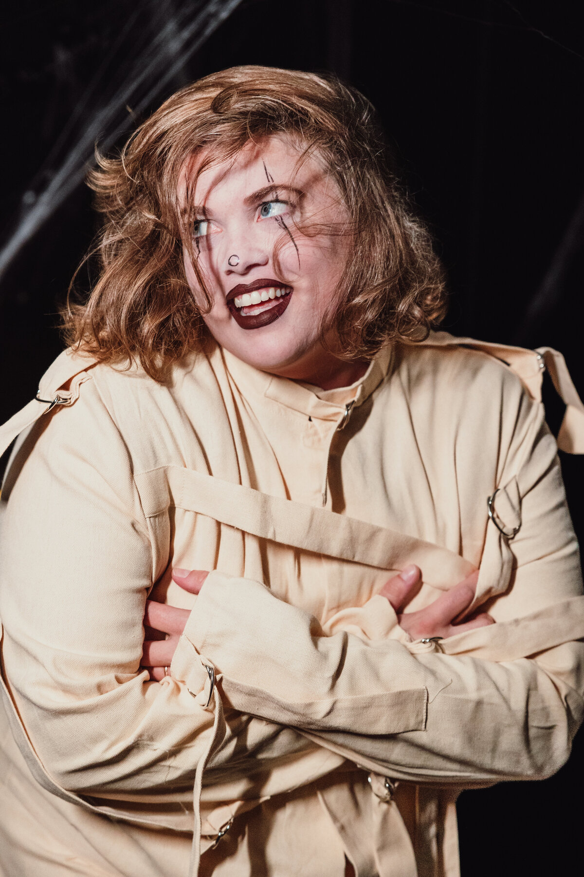Woman in straitjacket costume with face paint laughs dramatically in dark Halloween-themed boudoir in Tampa, Florida.
