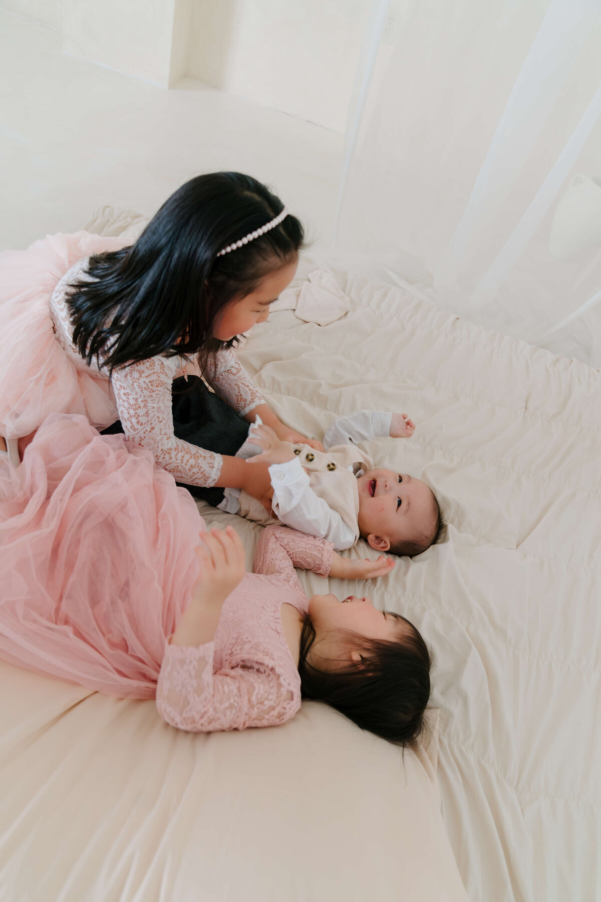 Two young girls in pink dresses happily laying on a bed with baby, enjoying a moment of play and friendship together.
