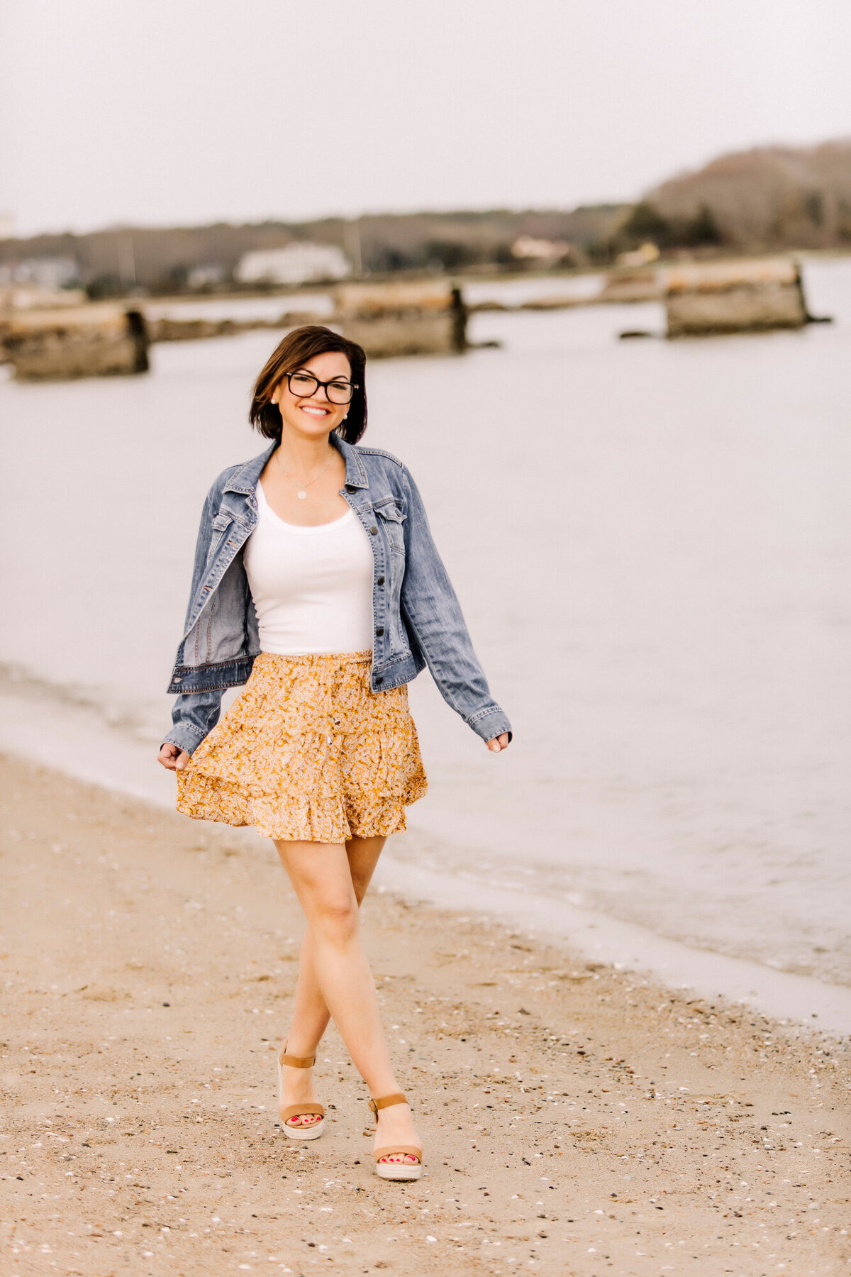A female dating portrait client wearing a jean jacket over a white shirt & flowy yellow skirt, walking down the beach