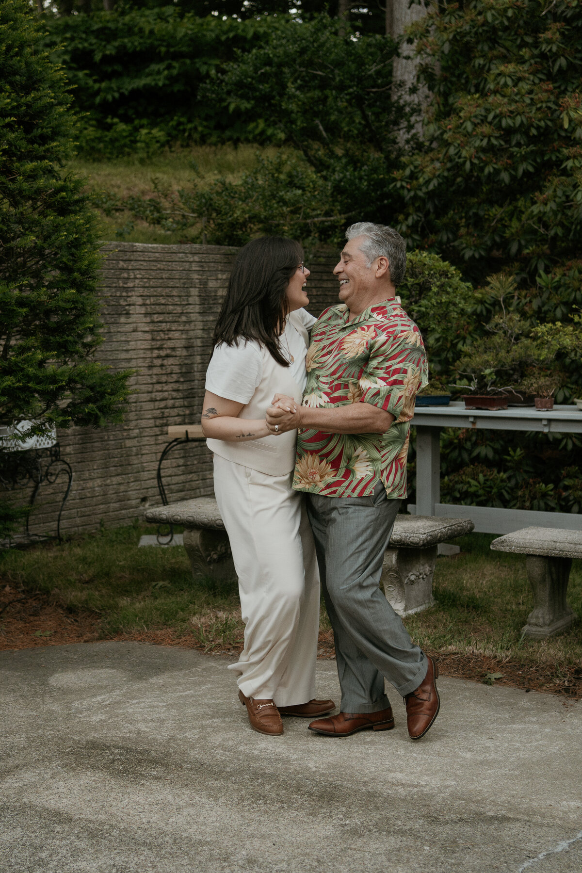 A joyful couple dances together outdoors, sharing a lighthearted moment. The woman, in a white outfit, and the man, in a colorful Hawaiian shirt and grey trousers, smile and laugh as they enjoy the dance. The lush greenery and stone benches in the background create a serene and relaxed atmosphere.