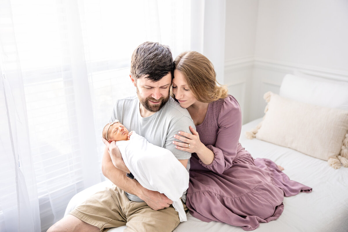 couple sitting on bed next to a window holding newborn baby