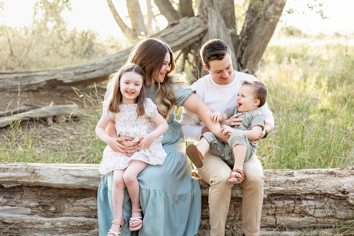 A family of four sitting on a fallen log in a wooded area, laughing and playing together.