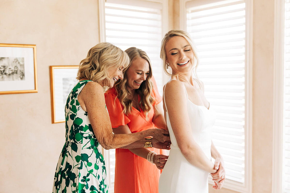 Two women smile while helping a bride zip her dress as she gets ready for her wedding planned by Perfect Thirds and Co. 