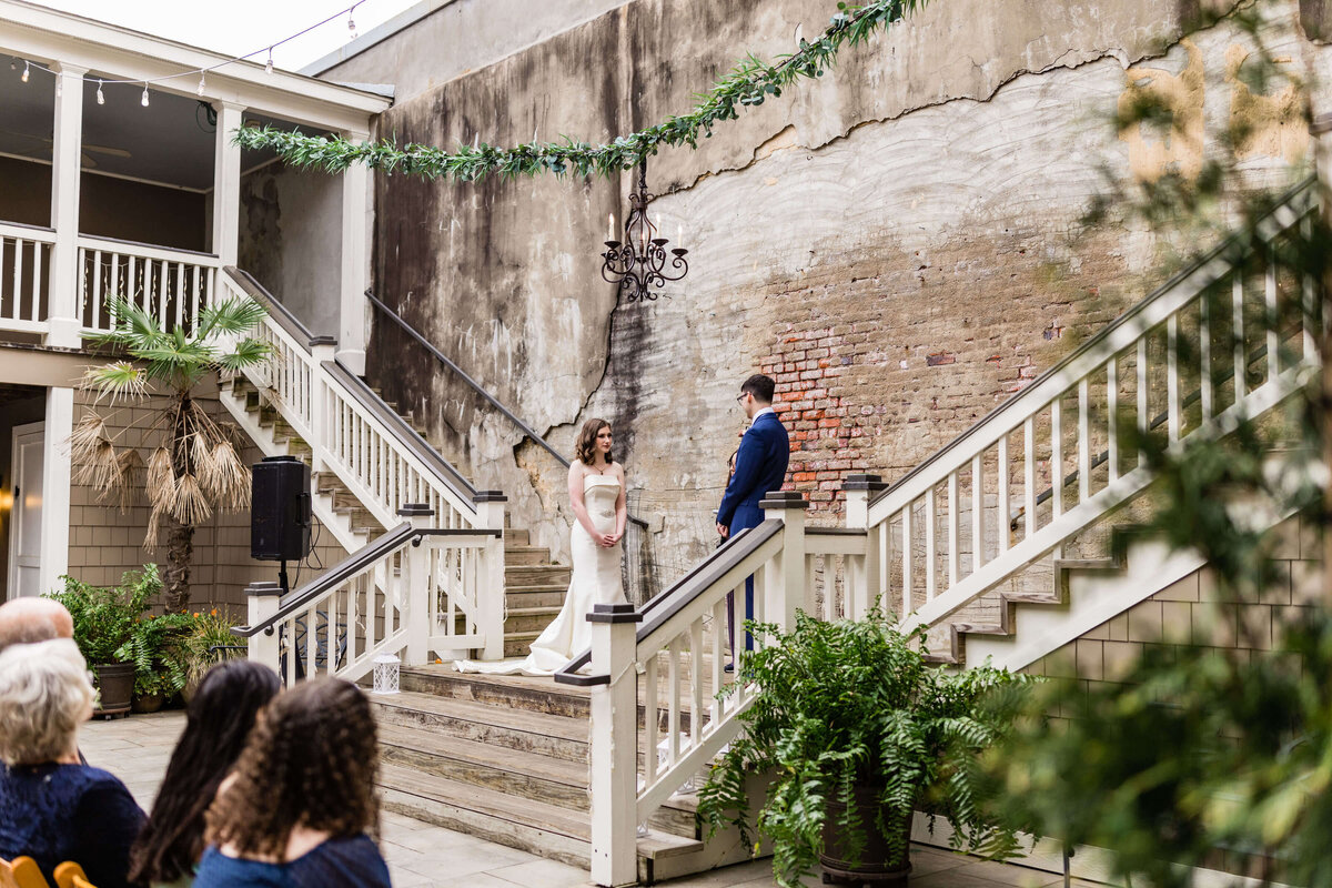 A bride and groom stand facing each other on wide steps in an outdoor courtyard with a brick wall, surrounded by greenery, as seated guests look on.