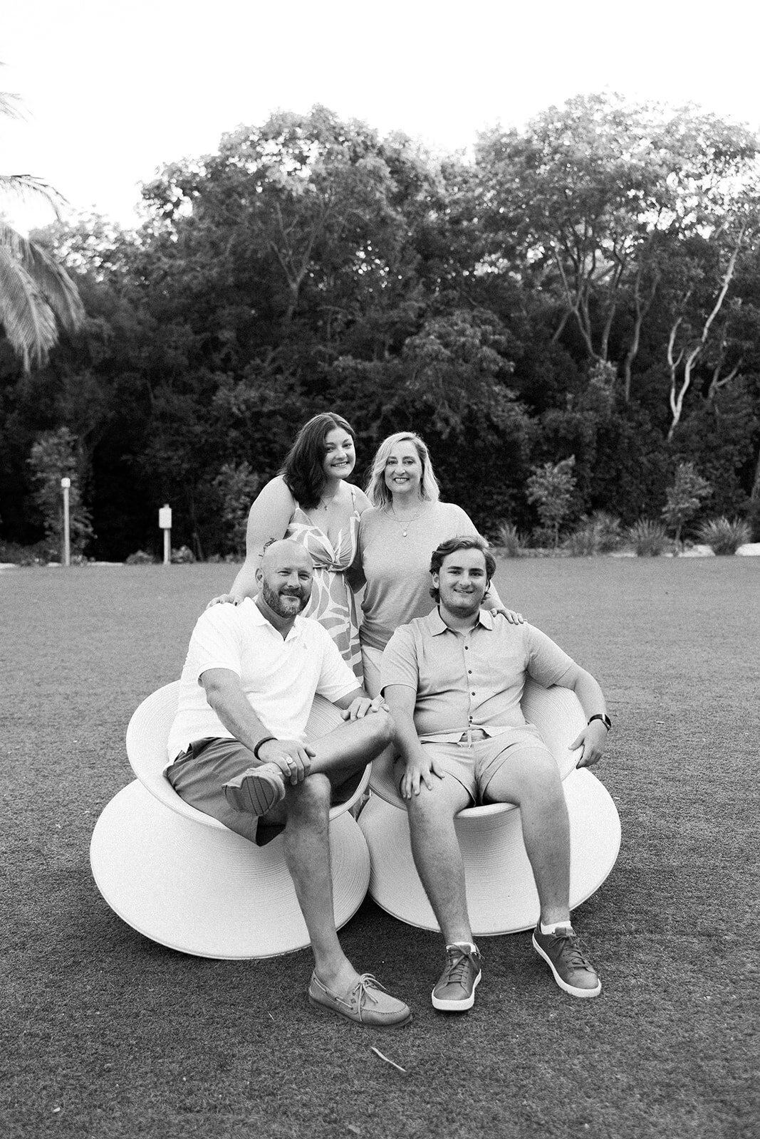 Black and white portrait of a family posing outdoors on modern white chairs, surrounded by trees. Florida Keys family portrait session by Claudia Amalia.