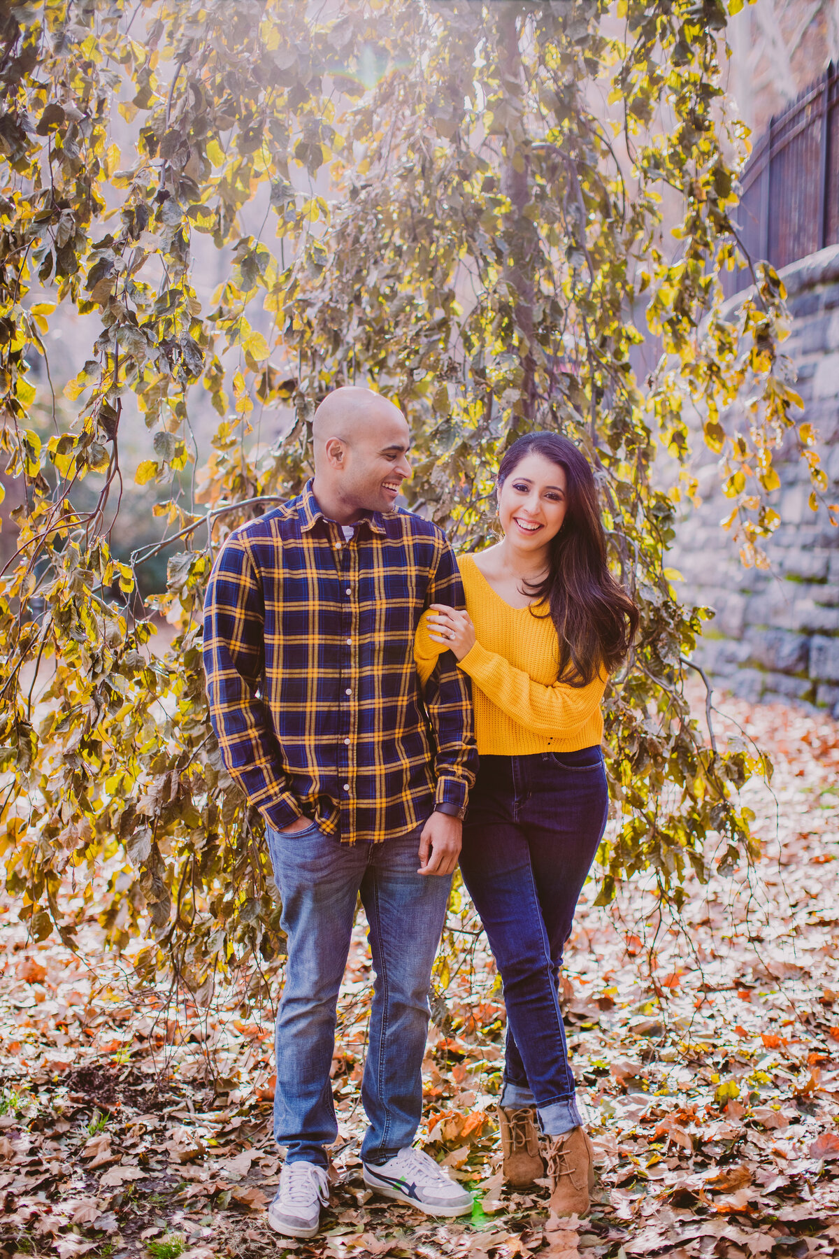 engaged couple in fall clothing stand in front of a tree at carl schurz park in nyc