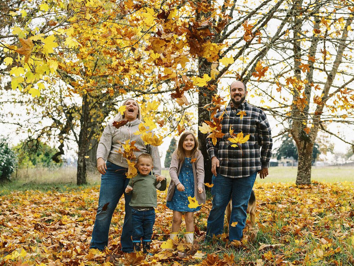 A family stands outdoors throwing leaves during a family photo shoot.