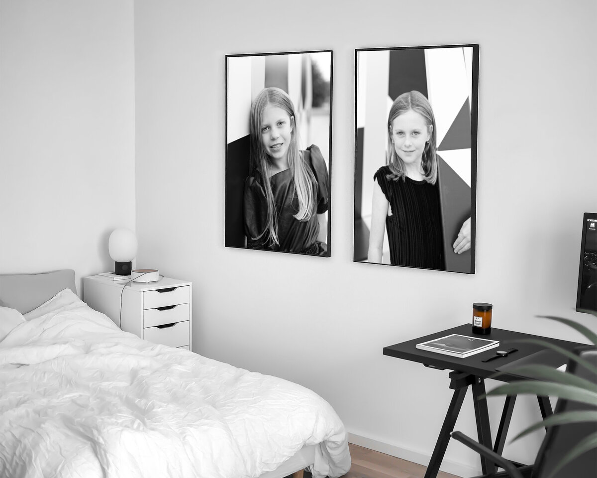 A modern bedroom with a neatly made bed, white side table, and a sleek black desk with a computer monitor. The wall features two large, framed black-and-white portraits of a young girl with long hair. A small potted plant sits on the desk.