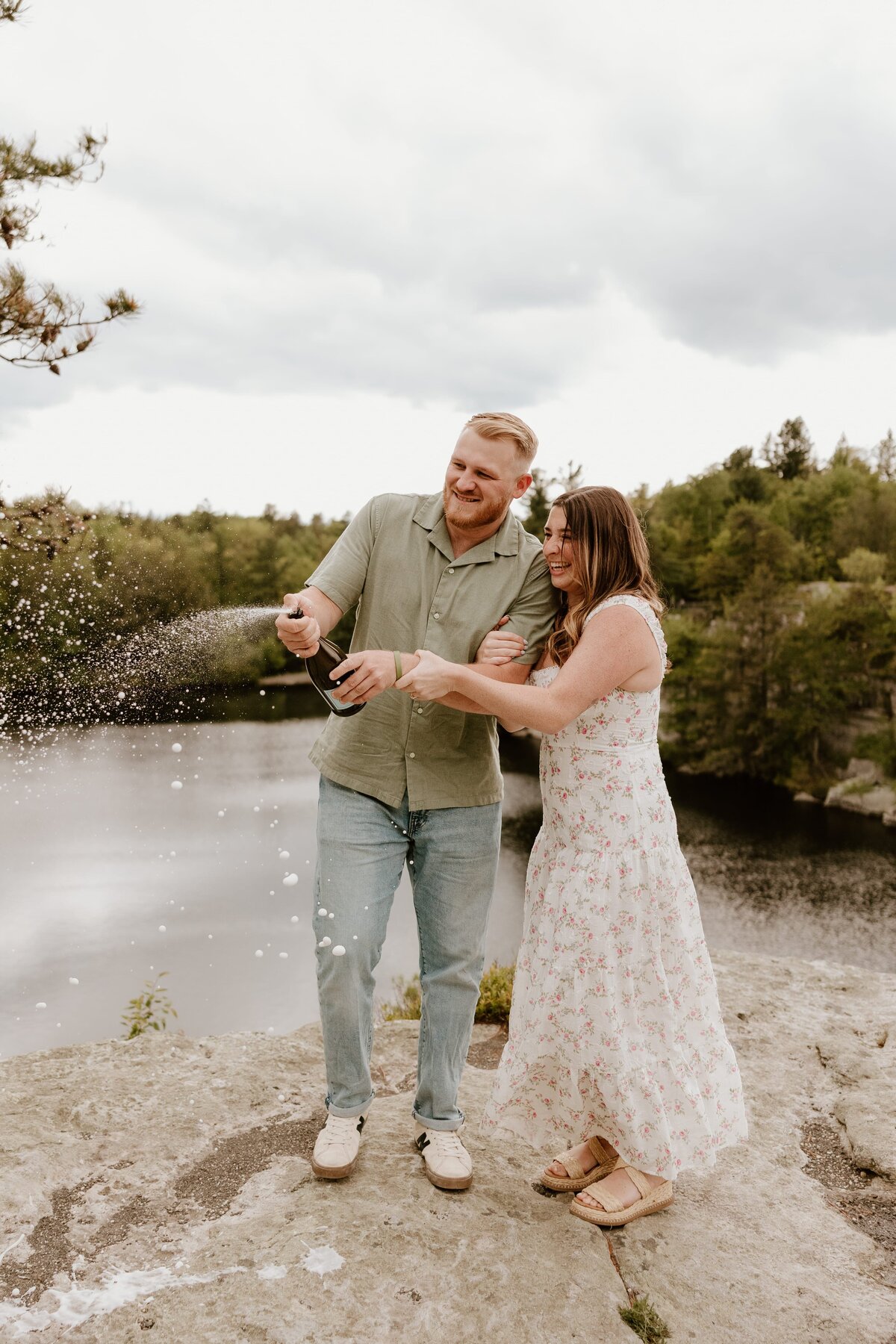 Engaged couple pops champagne together on a rocky cliffside overlooking a lake, laughing and celebrating with sparkling spray in the air.