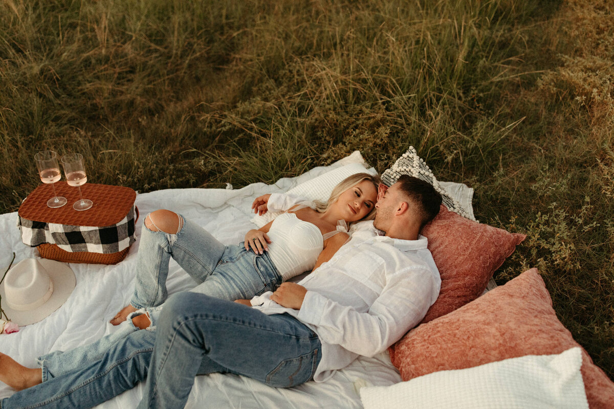 Couple laying down in a field on a blanket and pillows with a picnic basket and champagne glasses