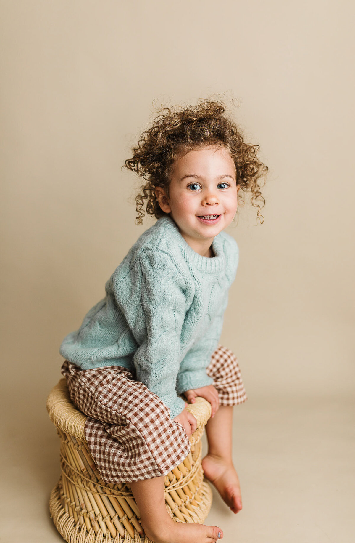 Toddler girl sitting on a rattan stool on a neutral beige studio backdrop, smiling 