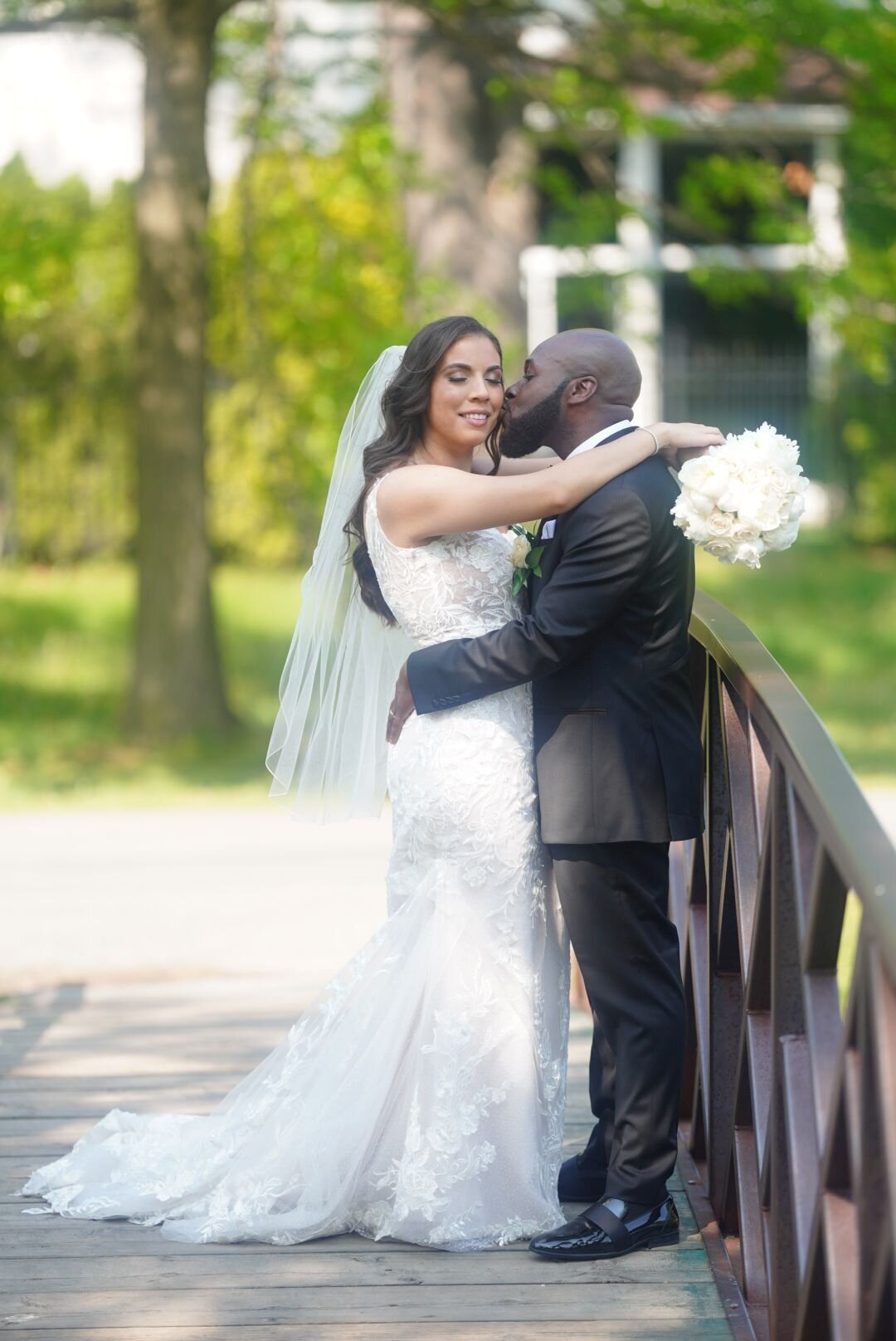 A couple stands on a scenic bridge in a nature park, embracing the serene surroundings and each other. The bridge and lush greenery create a picturesque setting, showcasing their romantic moment in a natural environment.