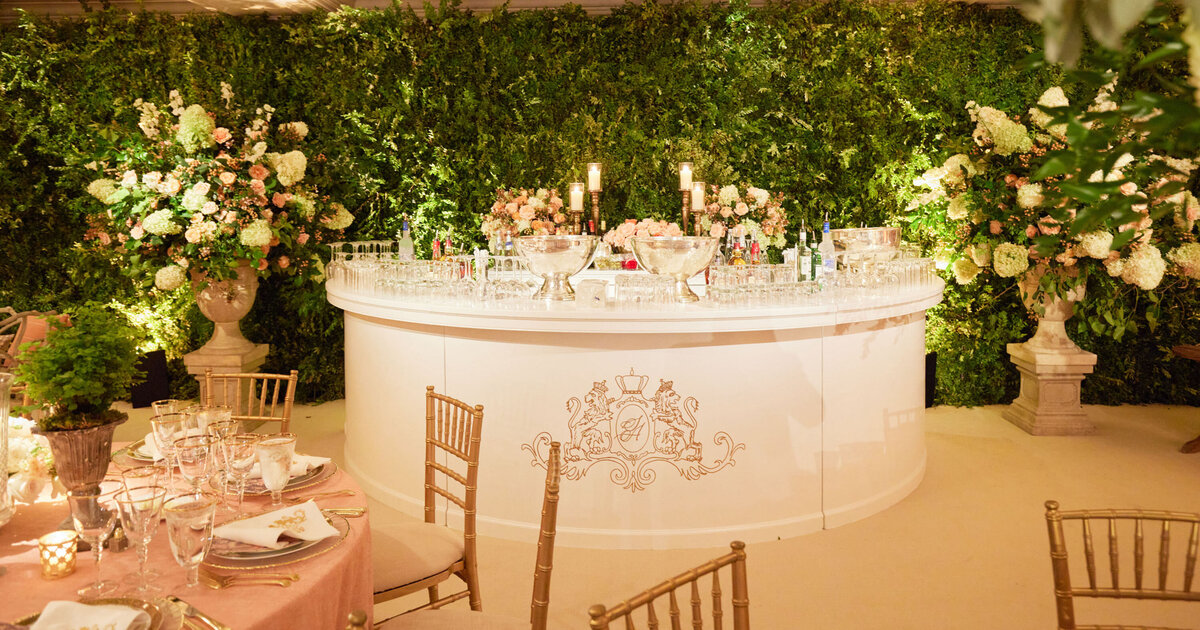 This captivating photo was taken at the prestigious Willard Intercontinental in Washington D.C., renowned as one of the top wedding venues and hotels in the capital city. Captured by the talented photographer Shawn Connell, the picture showcases a luxurious event setup. At the centerpiece stands a magnificent bar, exquisitely embellished with an ornate crest and encircled by verdant greenery and opulent floral arrangements. The bar itself is tastefully adorned with sparkling glassware, flickering candles, and intricate decorations, adding a touch of glamour to the setting. In the foreground, a charming table draped in a soft pink tablecloth is paired with elegant gold chairs, creating a sophisticated dining arrangement. Additionally, Clayton Films can be credited as the Videographers who beautifully captured this memorable event setup.