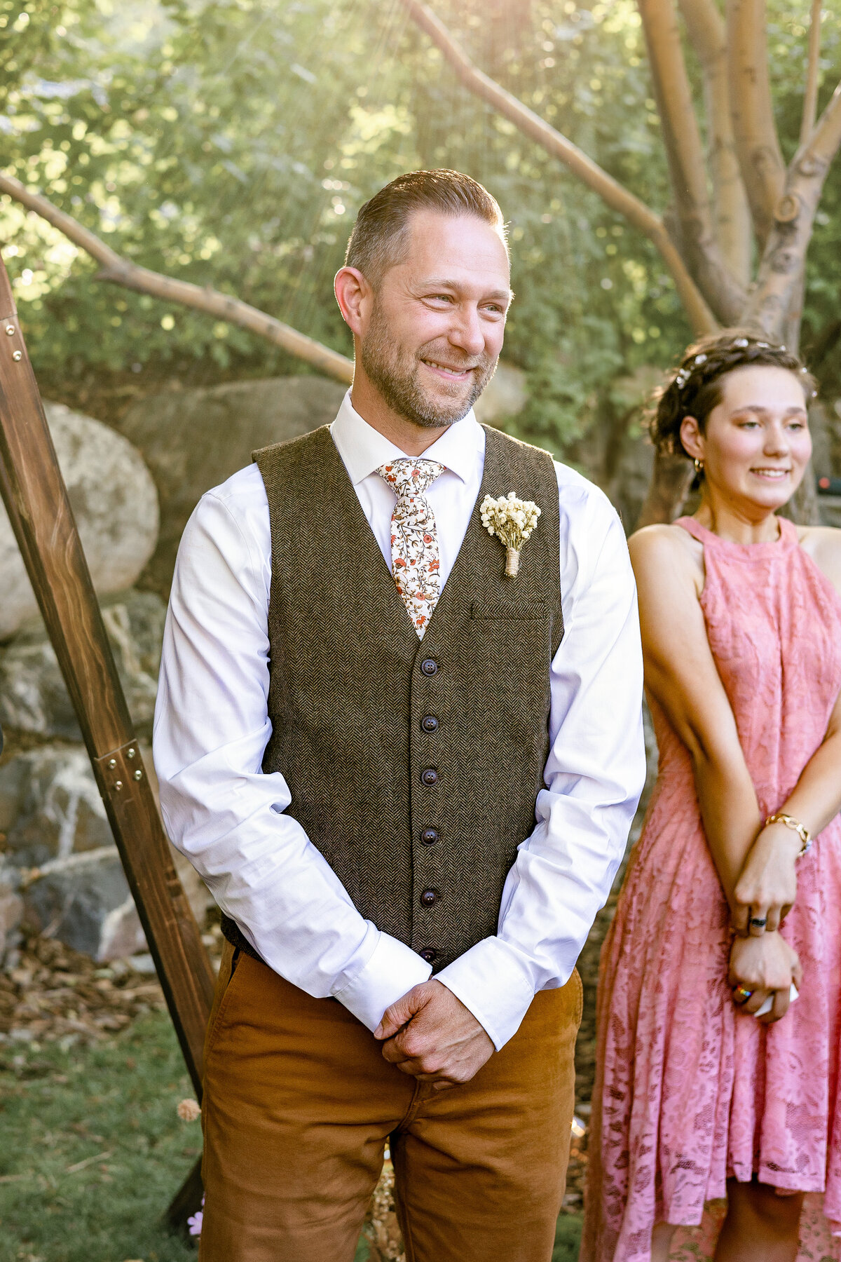 The groom stands smiling under dappled sunlight in Provo Canyon, wearing a floral tie, white shirt, and brown vest with a boutonniere, with tears in his eyes as he sees his bride for the first time.