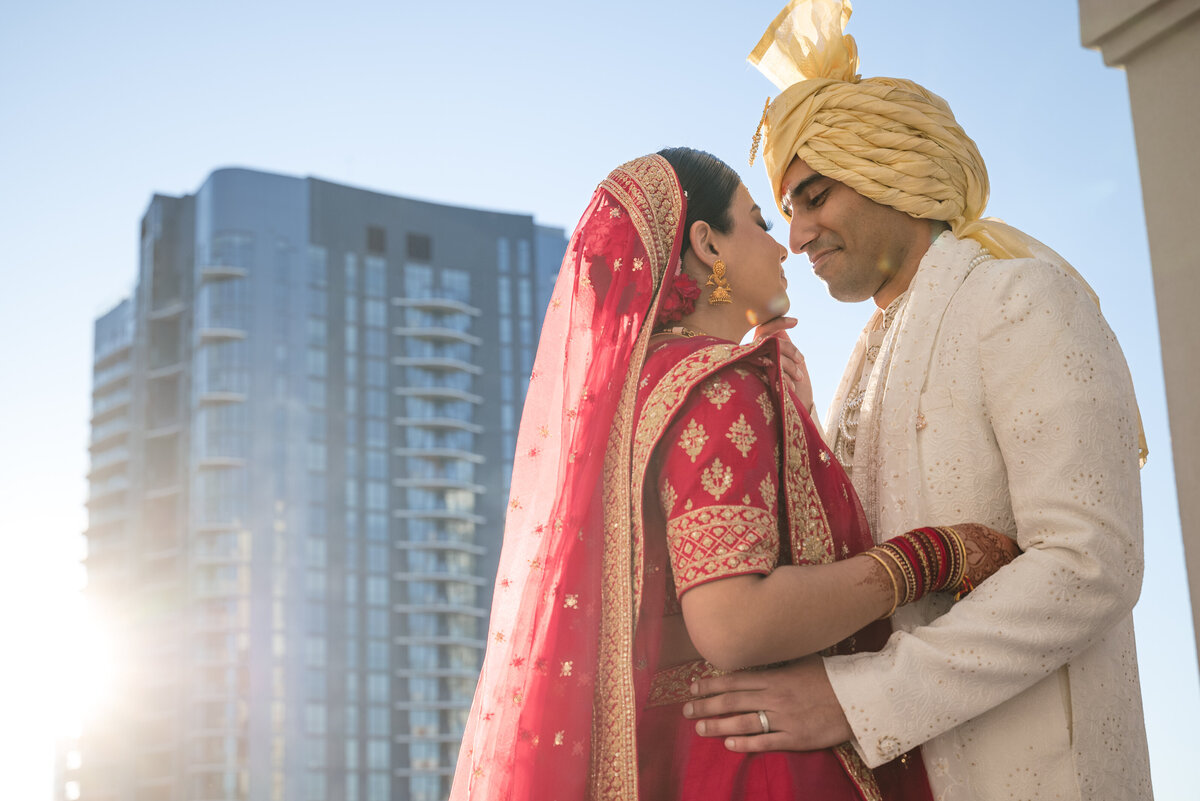 Romantic Indian wedding couple portrait at the Grand Bohemian Hotel Charlotte. Bride in red lehenga with gold jewelry and groom in ivory sherwani with golden turban share an intimate moment on the rooftop. Captured by DeLong Photography, Charlotte Indian wedding photographers specializing in cultural celebrations.