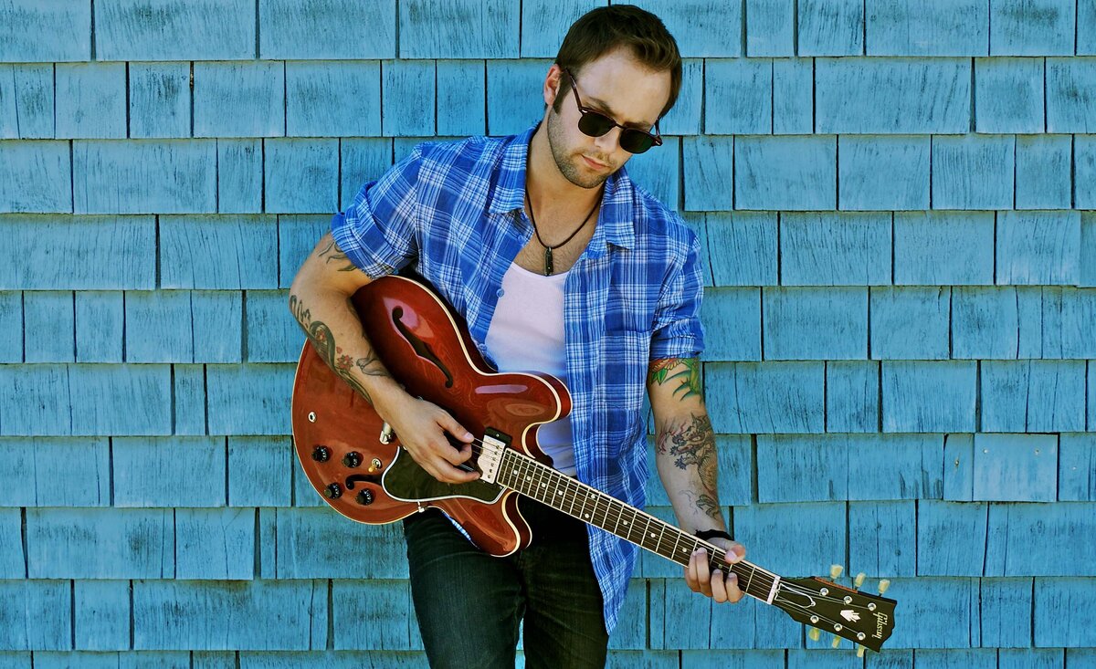 Male musician photo Dallas Smith wearing blue plaid shirt with sunglasses playing red electric guitar leaning against pale blue wood wall