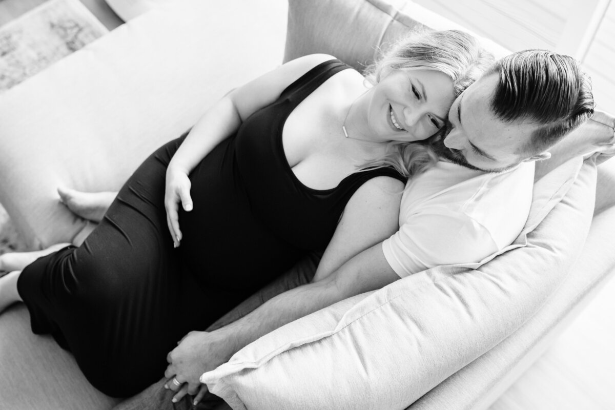 A pregnant woman in a black dress and her partner lie close together on a couch, the woman resting her head on the man's chest while they smile warmly at each other.