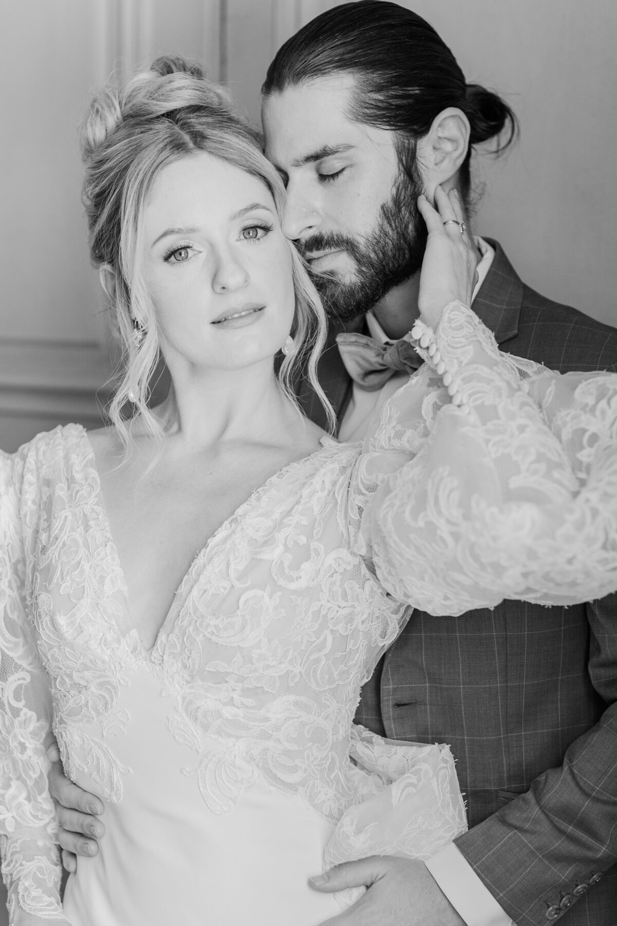 Black and white image. Close up of bride and groom. The bride's back is to the groom, her arm reaching up and gently touching his neck. The groom's eyes are closed and is resting forehead against the bride's temple. The bride is looking at the camera with a half smile. Editorial feel. Captured by Paris Destination Wedding Photographer Lia Rose Weddings